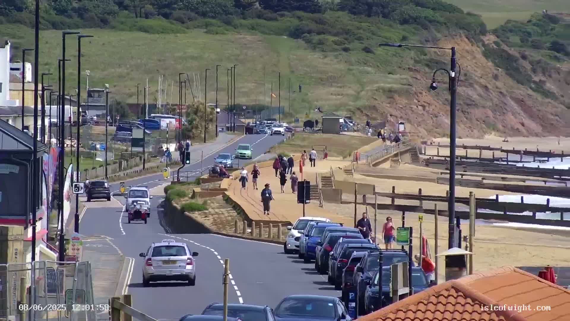 A sunny seaside road scene shows cars parked along a sandy beach with pedestrians walking on a nearby paved path.