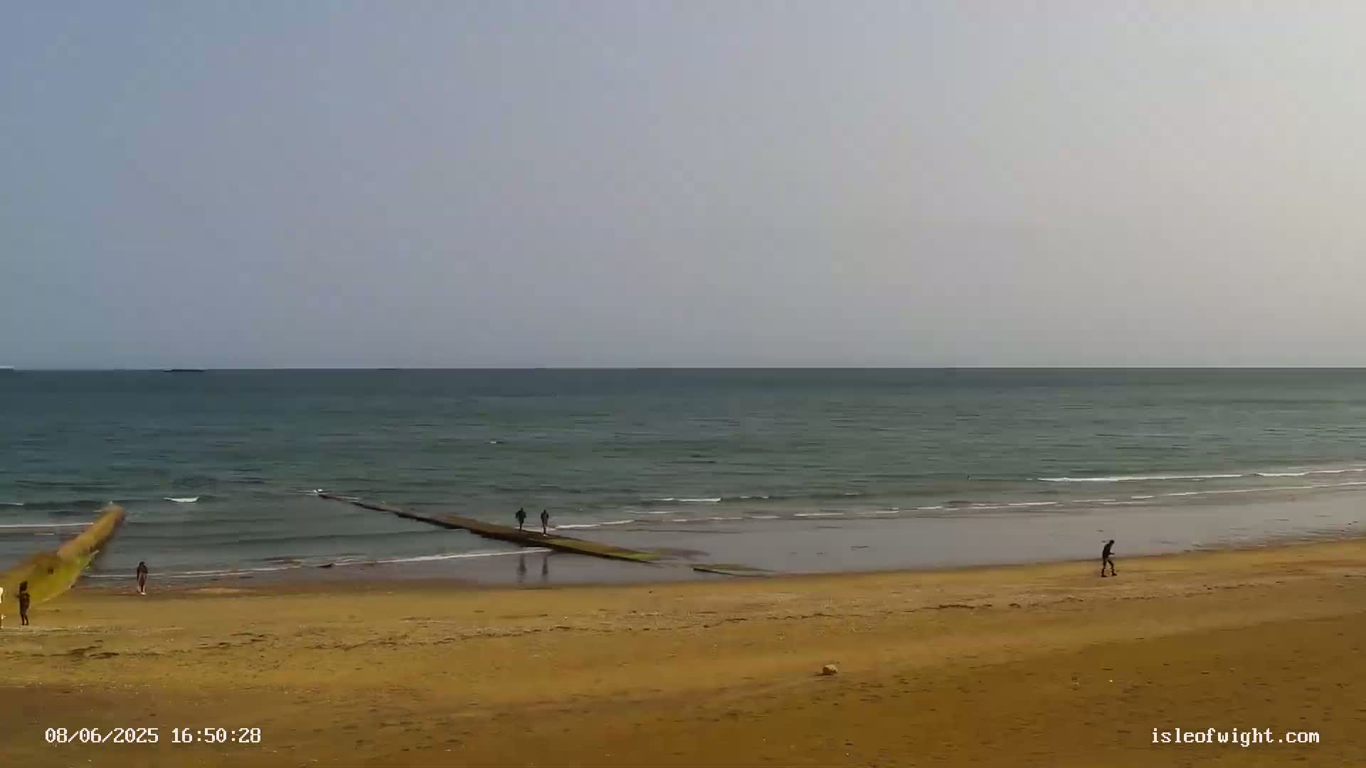 A mostly calm sea meets a sandy beach where several people walk along the shore and a short jetty.