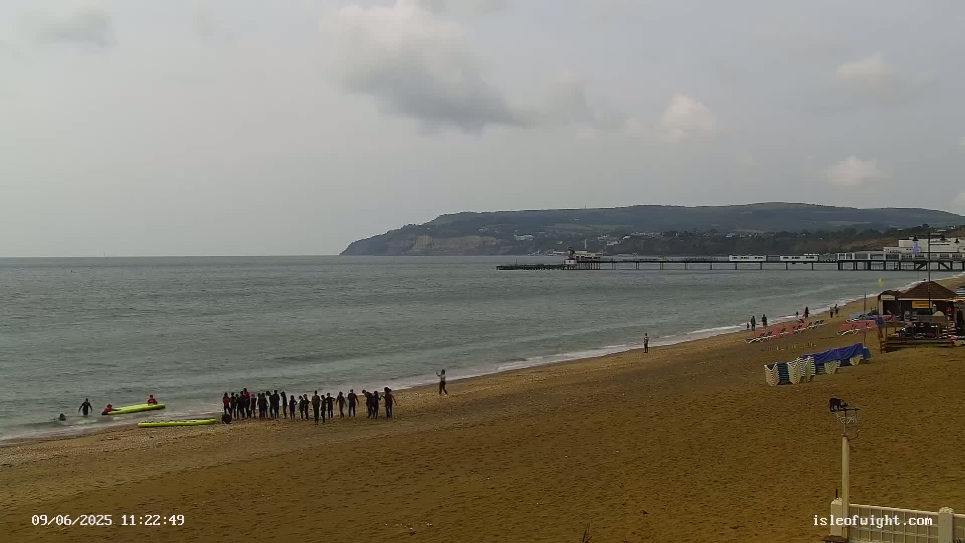 A sandy beach with a group of people standing near the water's edge, a pier extending into a calm sea under an overcast sky, and a distant cliff in the background.