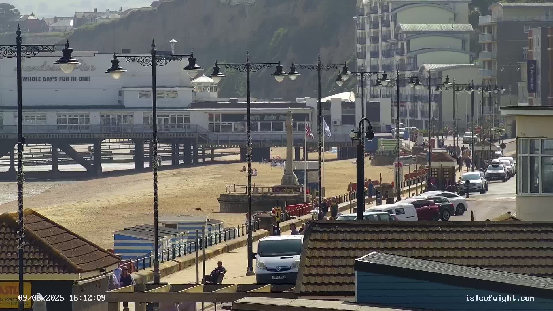 A pier, beach, and boardwalk are visible on a sunny day, with cars parked along the road and people walking on the boardwalk.