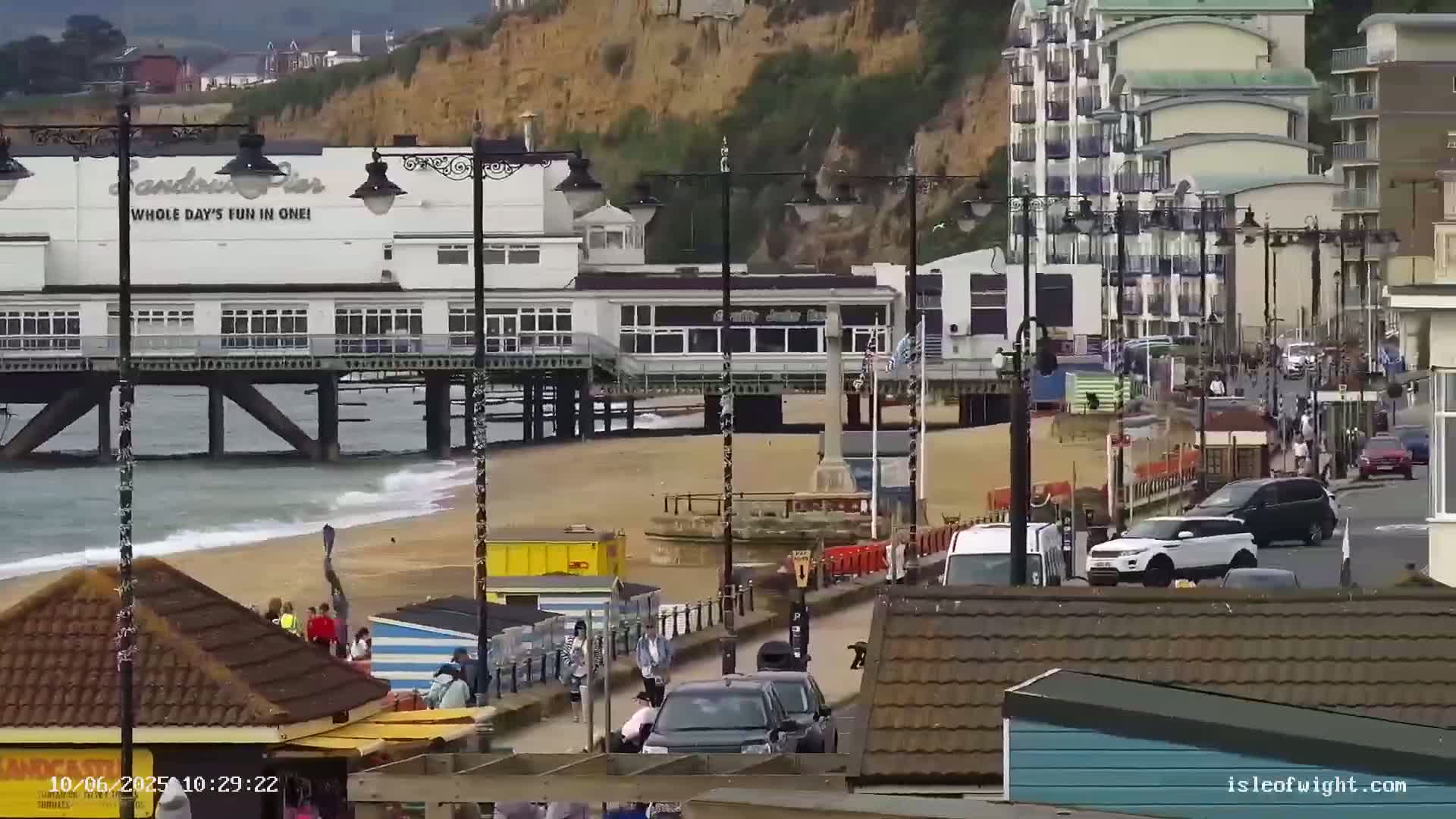 A pier extends over a sandy beach with gentle waves, backed by buildings and a cliff face, under overcast skies.