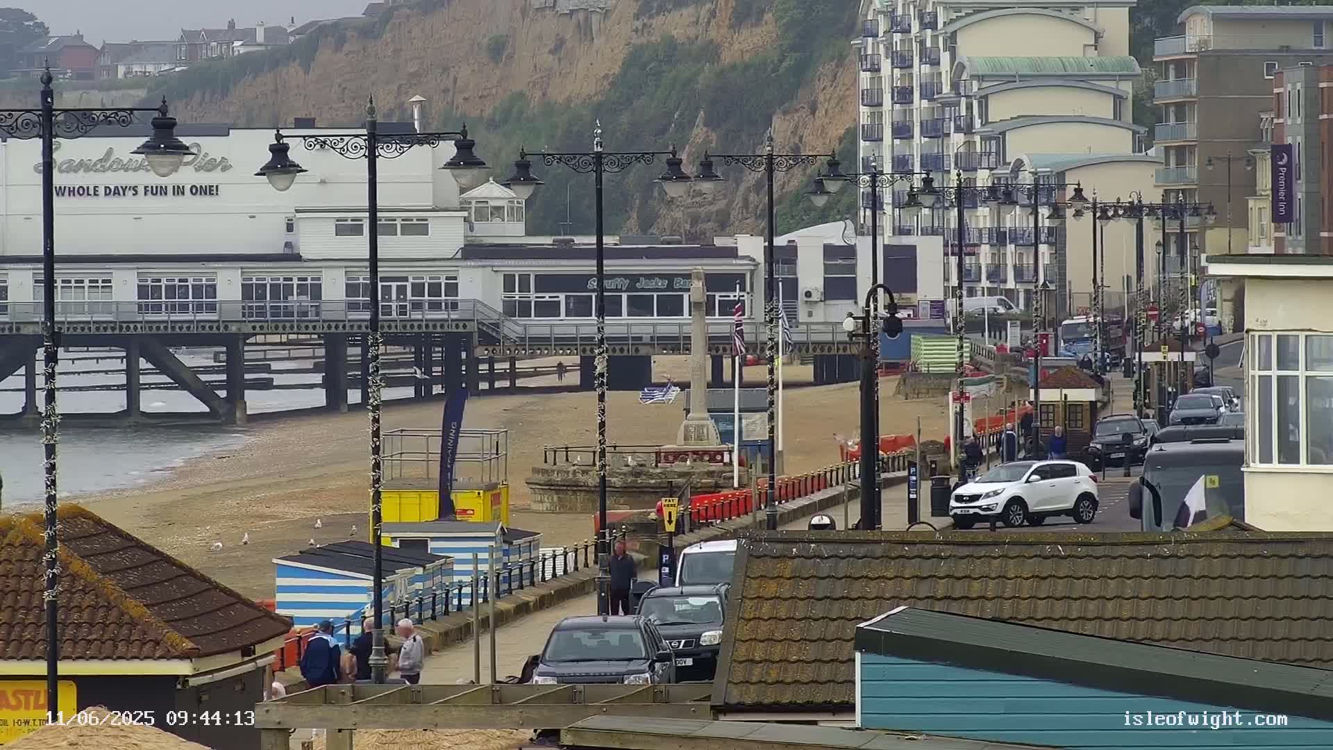 A seaside pier with buildings and cars on a cloudy day, with a sandy beach in the foreground.