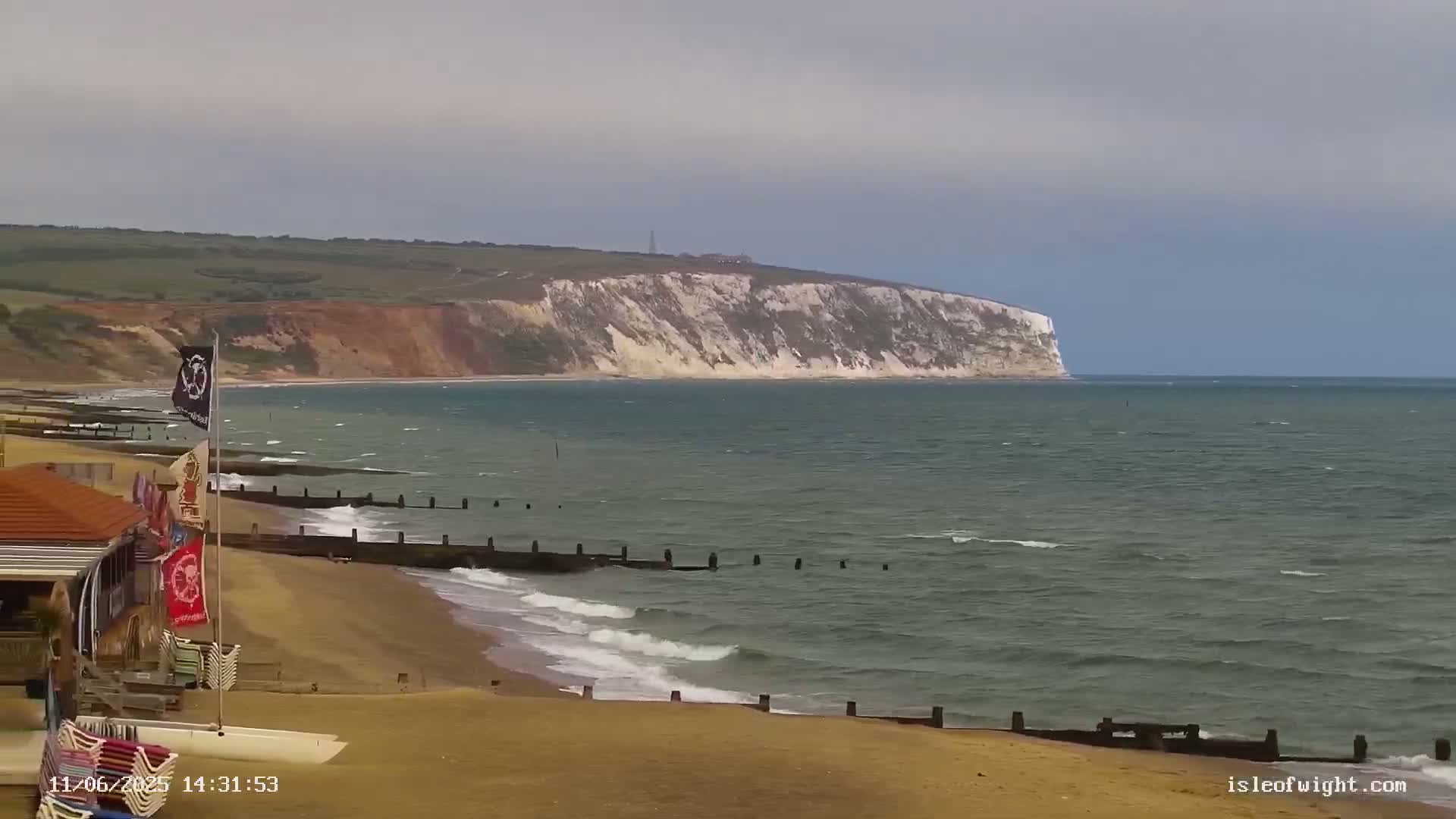 A sandy beach with a small building and flags stretches toward a calm sea meeting a large white cliff under an overcast sky.
