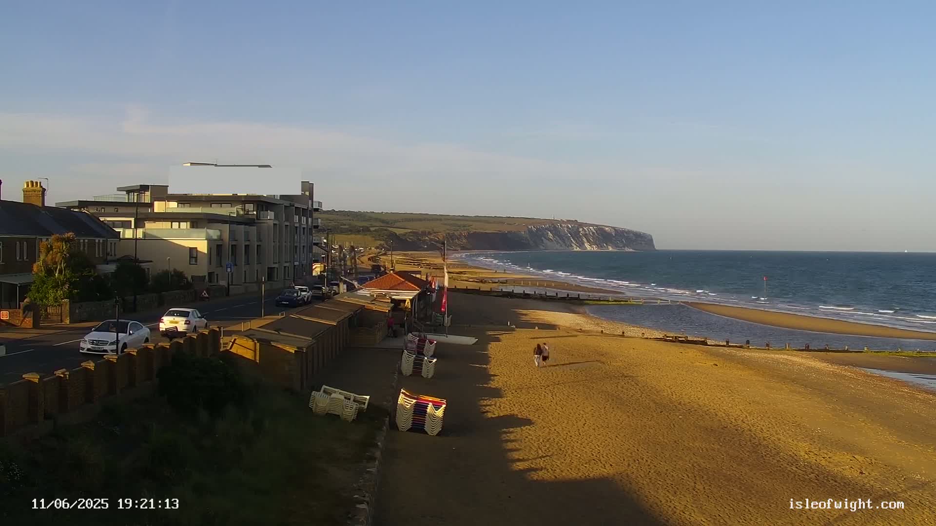 A sandy beach with a few people walking on it stretches towards a calm ocean and a distant cliff under a clear sky.