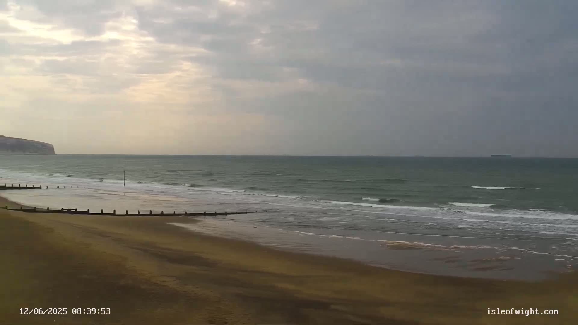 A sandy beach with gentle waves rolls onto the shore under a cloudy sky, with a distant cliff visible to the left.