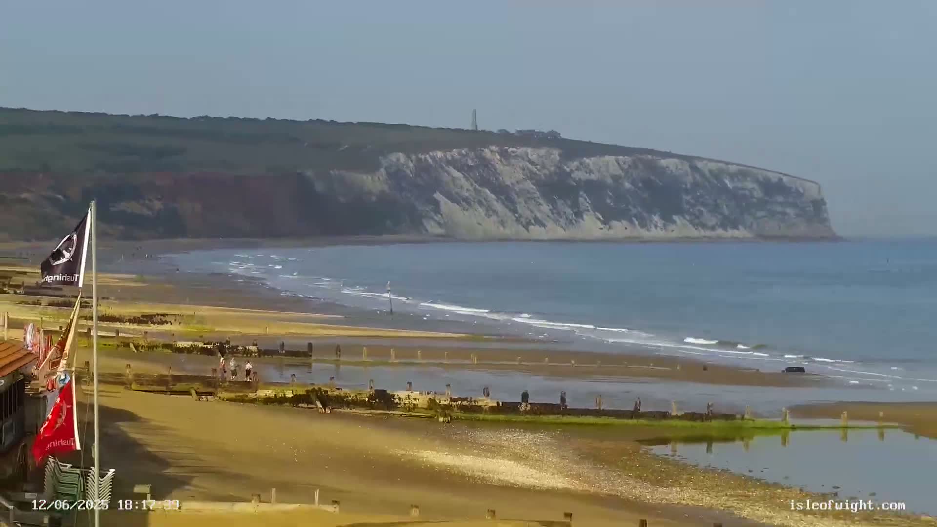 A sandy beach with gentle waves stretches towards a large, white cliff face under a hazy sky.