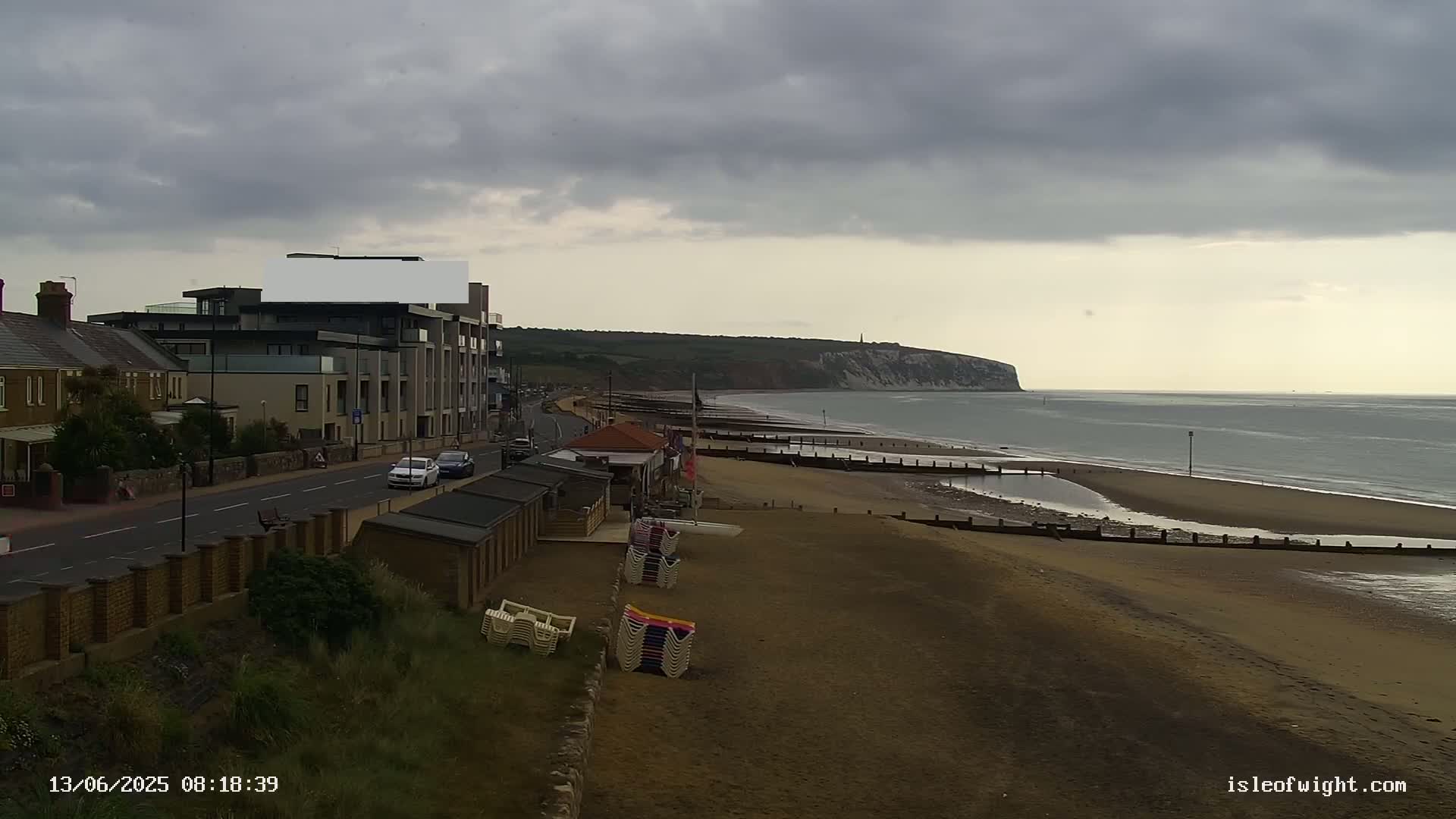 A sandy beach with stacked beach chairs stretches out to a calm sea under a cloudy sky, with buildings and a road visible in the foreground.
