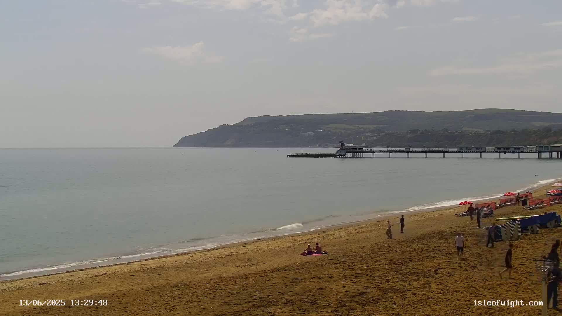 A sandy beach with a few people on it stretches to a calm sea, beyond which a pier and a distant hill are visible under a partly cloudy sky.