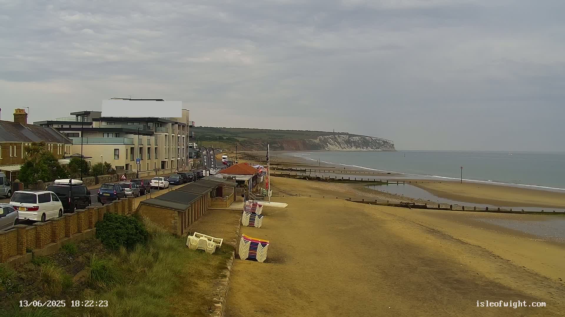 A sandy beach with stacked beach chairs stretches along a calm ocean toward a distant white cliff under an overcast sky, with a road and buildings visible in the foreground.