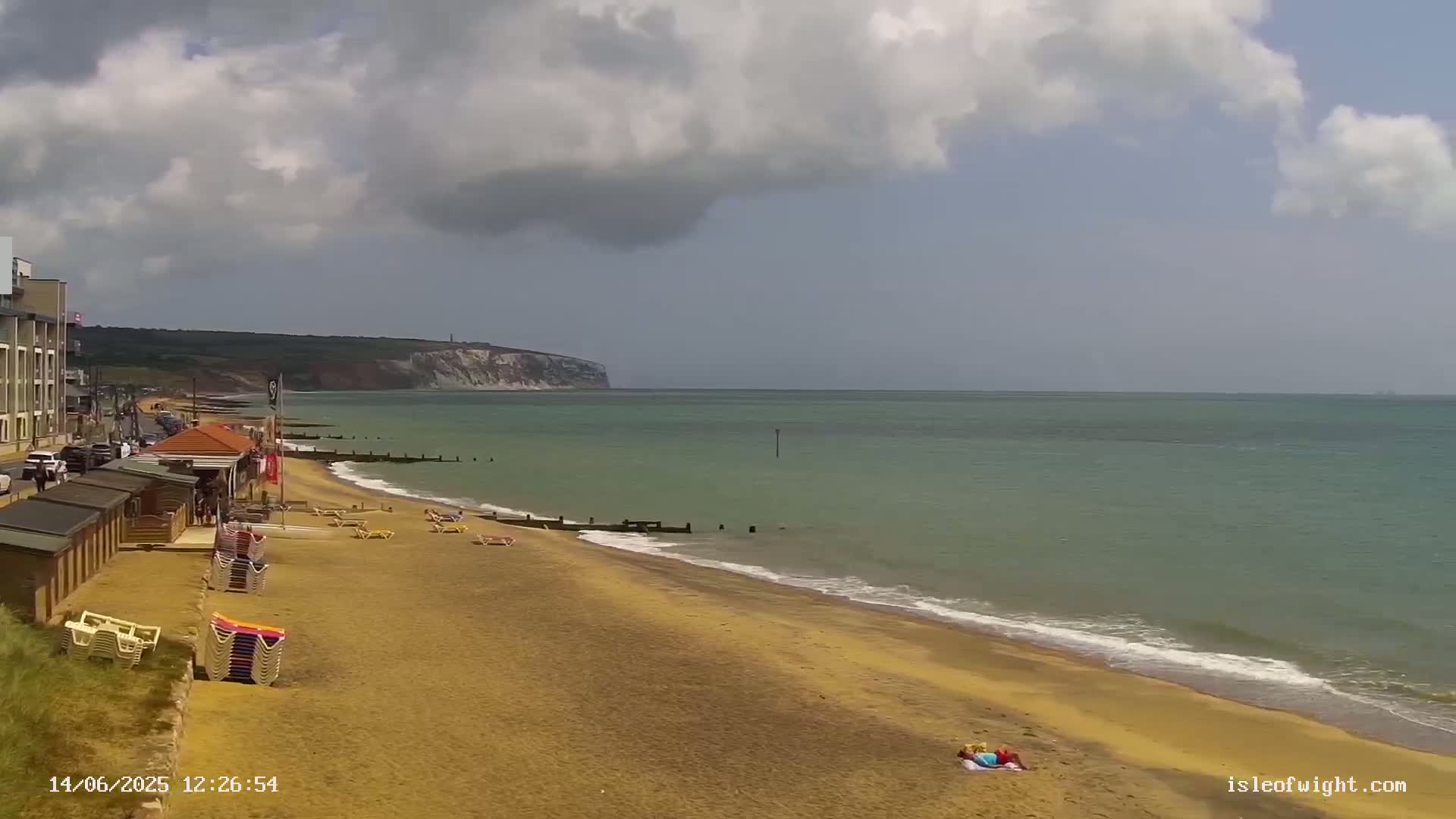 A sandy beach with beach huts and sunbathers stretches towards a calm sea under a partly cloudy sky, with a distant cliff face visible.