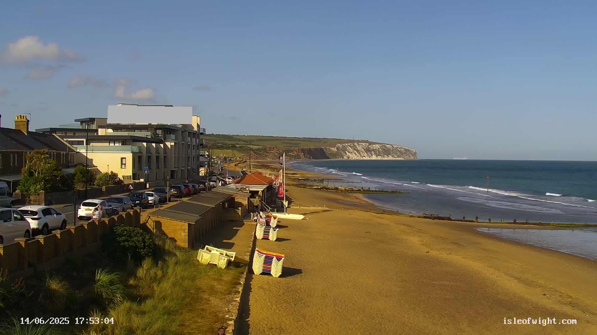 A sandy beach with small beach huts and a building lines the coast, under a clear sky, with gentle waves lapping the shore and a distant cliff visible.