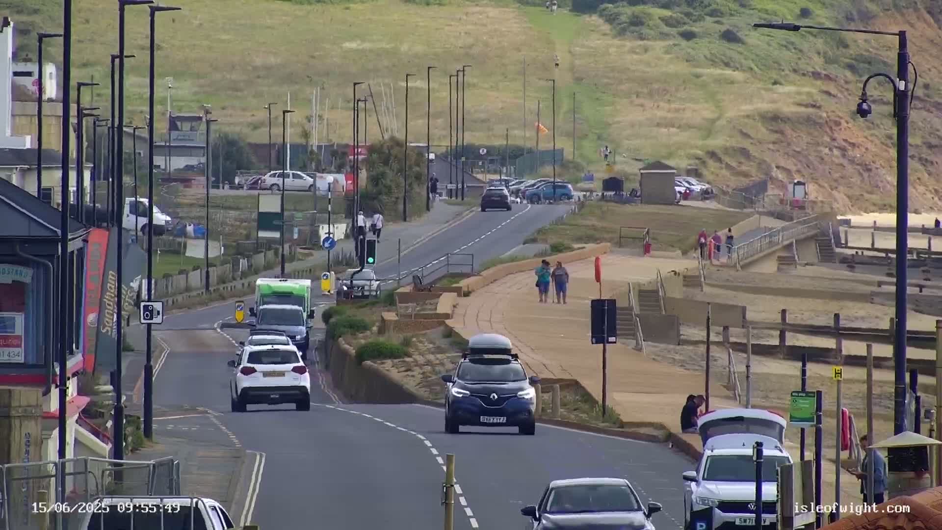 A coastal road with several cars driving on it, pedestrians walking along a nearby sandy beach, and a partly cloudy sky.