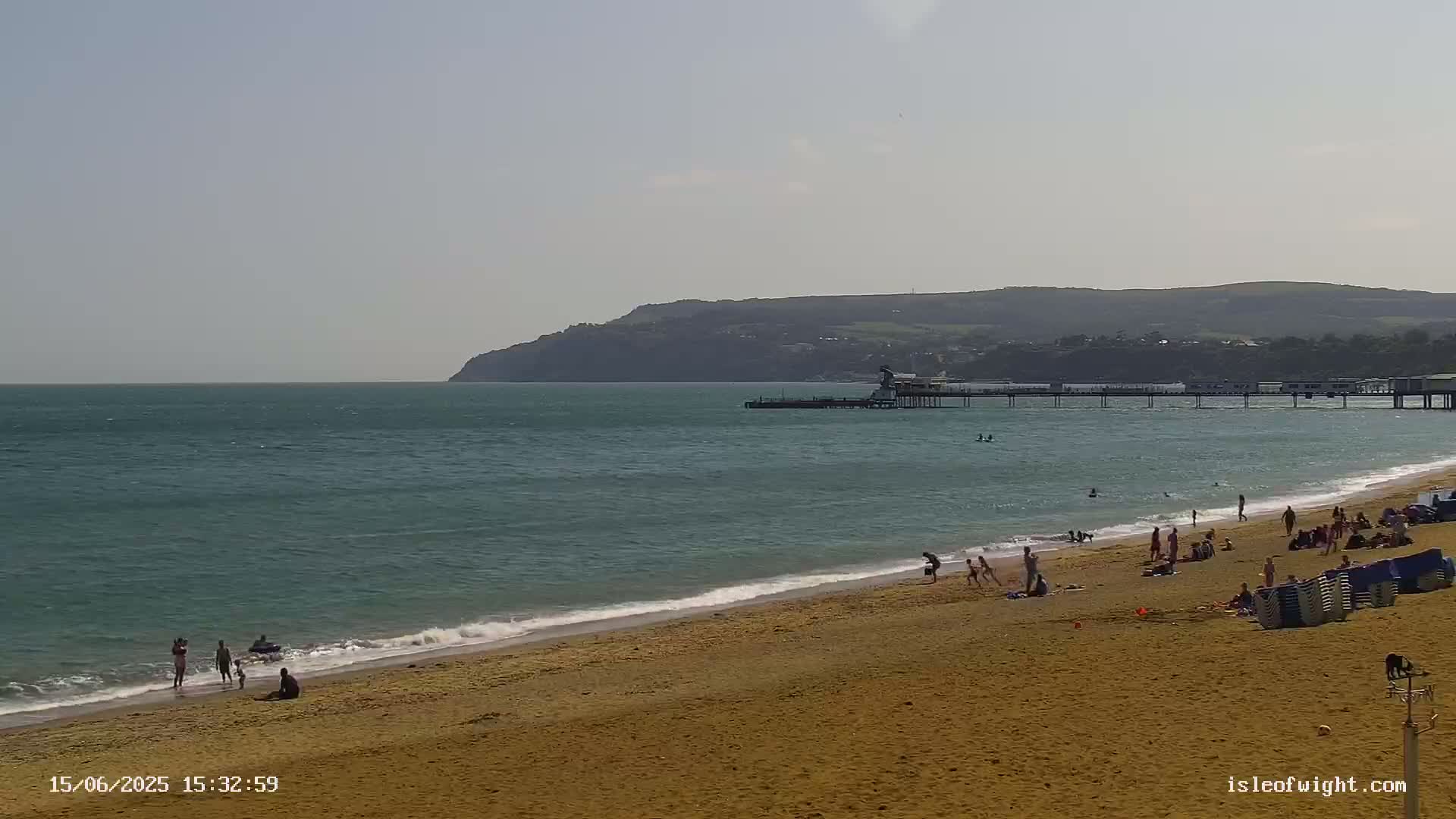 A sandy beach with scattered people and a pier extends into a calm, teal sea under a bright, clear sky, with a distant hill visible in the background.
