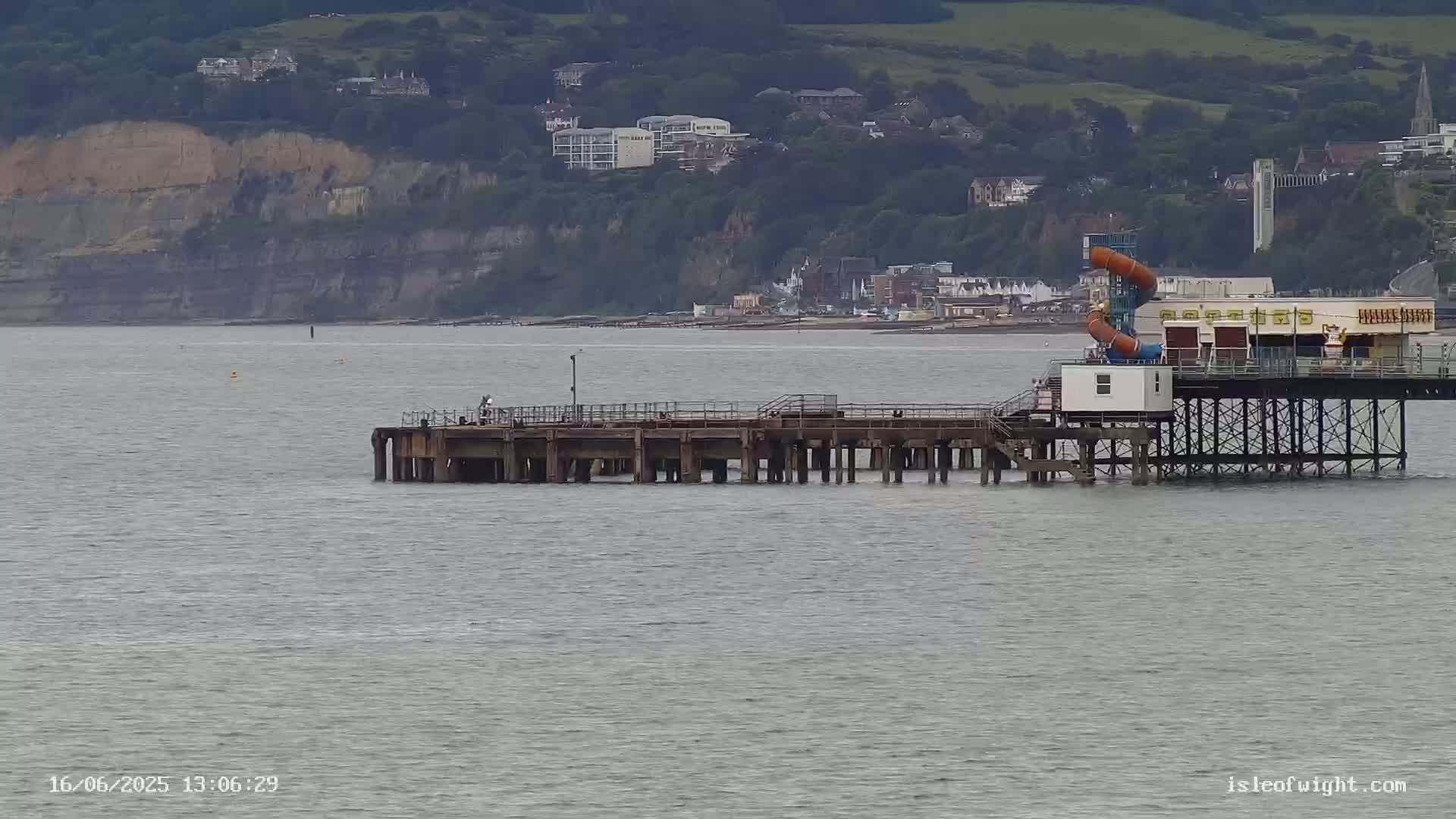 A pier with a water slide extends into a calm body of water, with a cliff and town visible in the background.