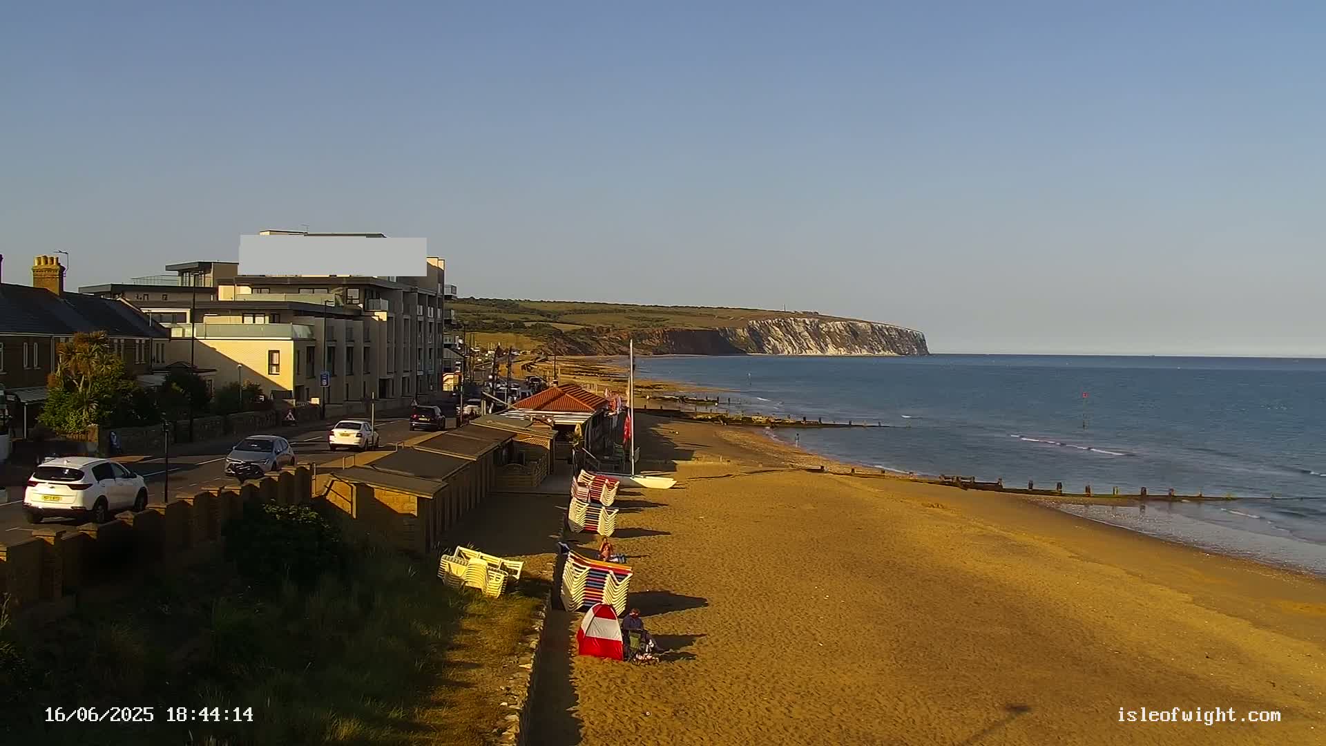 A sandy beach with beach huts and deck chairs stretches towards a calm sea and white cliffs under a clear sky.