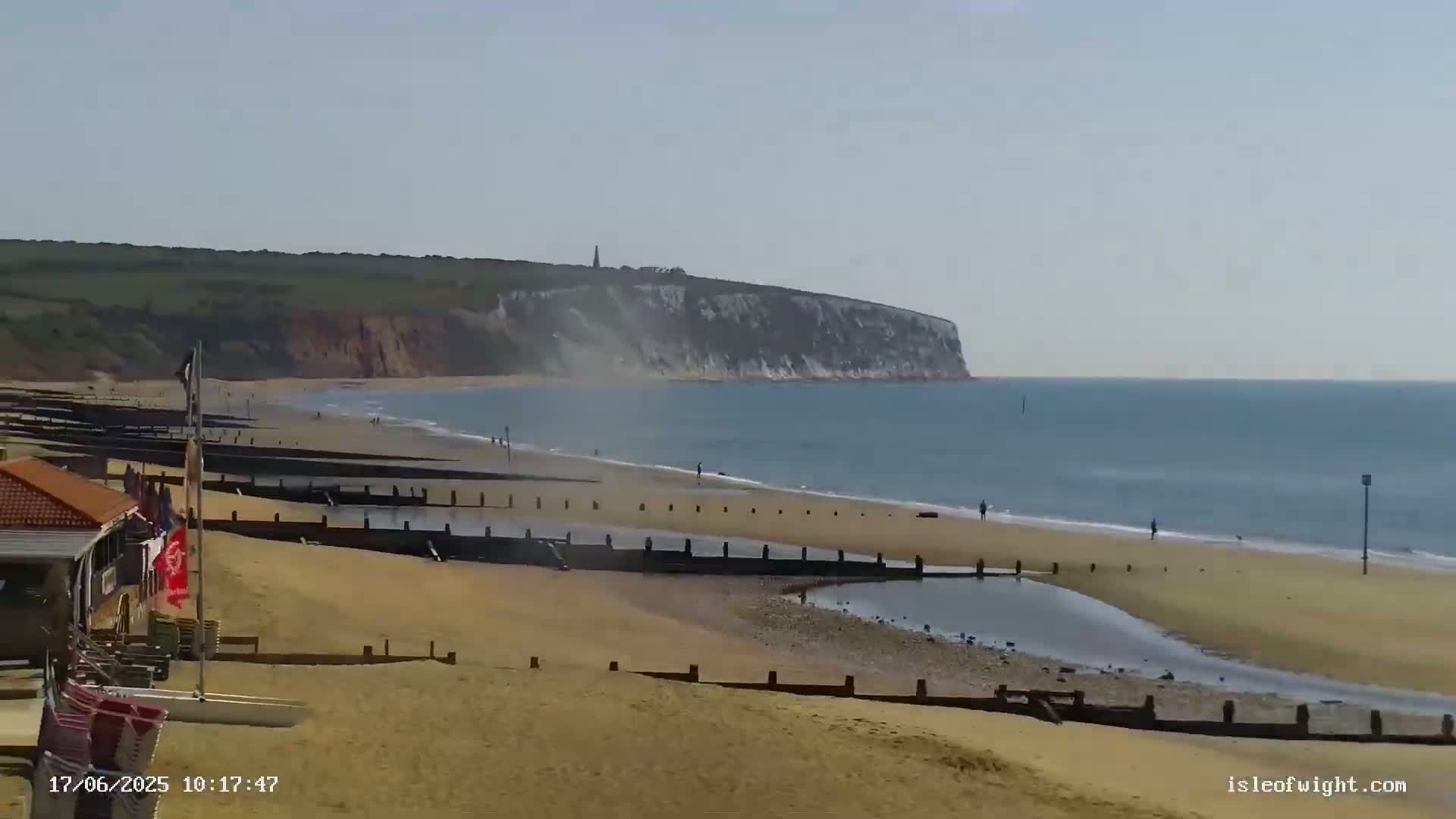 A sandy beach with wooden posts and a small body of water stretches towards a large, light-colored cliff in the distance under a mostly clear sky.