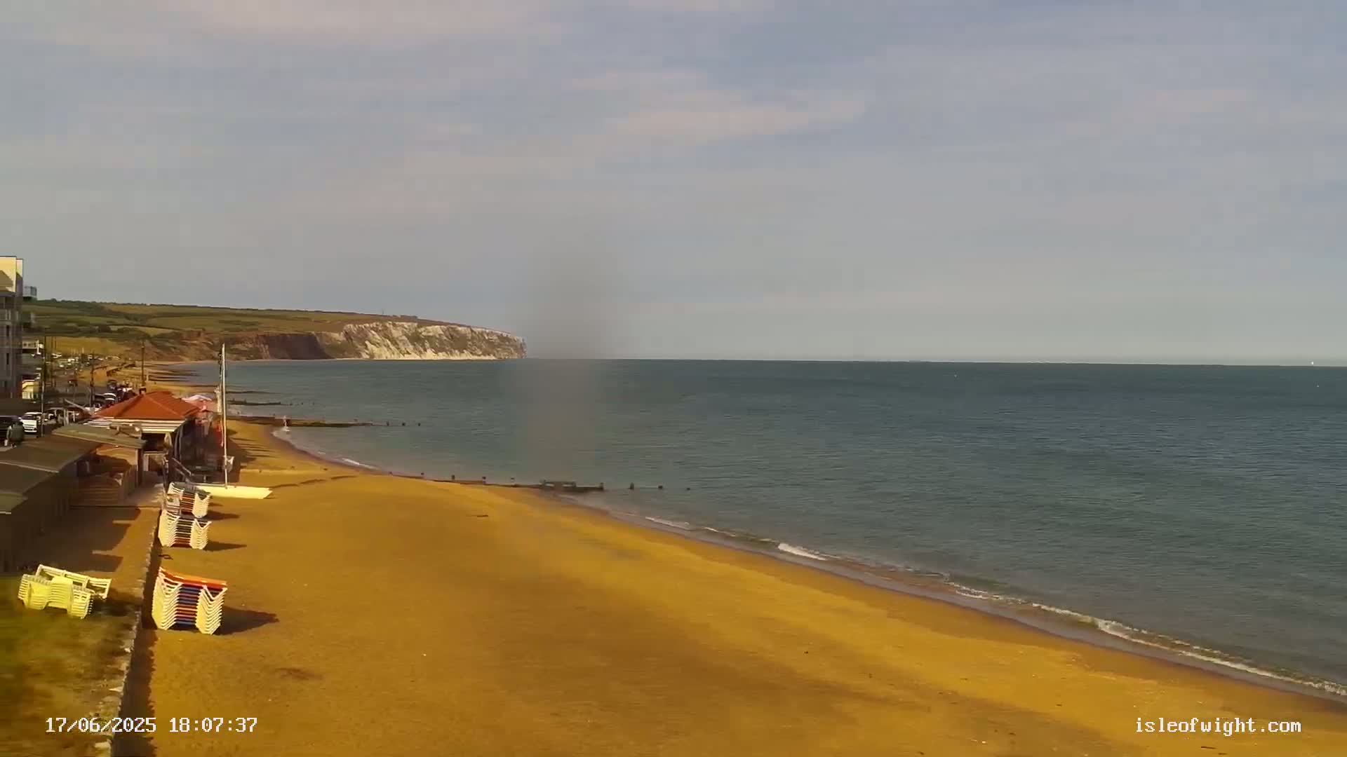A mostly sunny day shows a sandy beach with stacked beach chairs next to a calm sea and distant white cliffs.