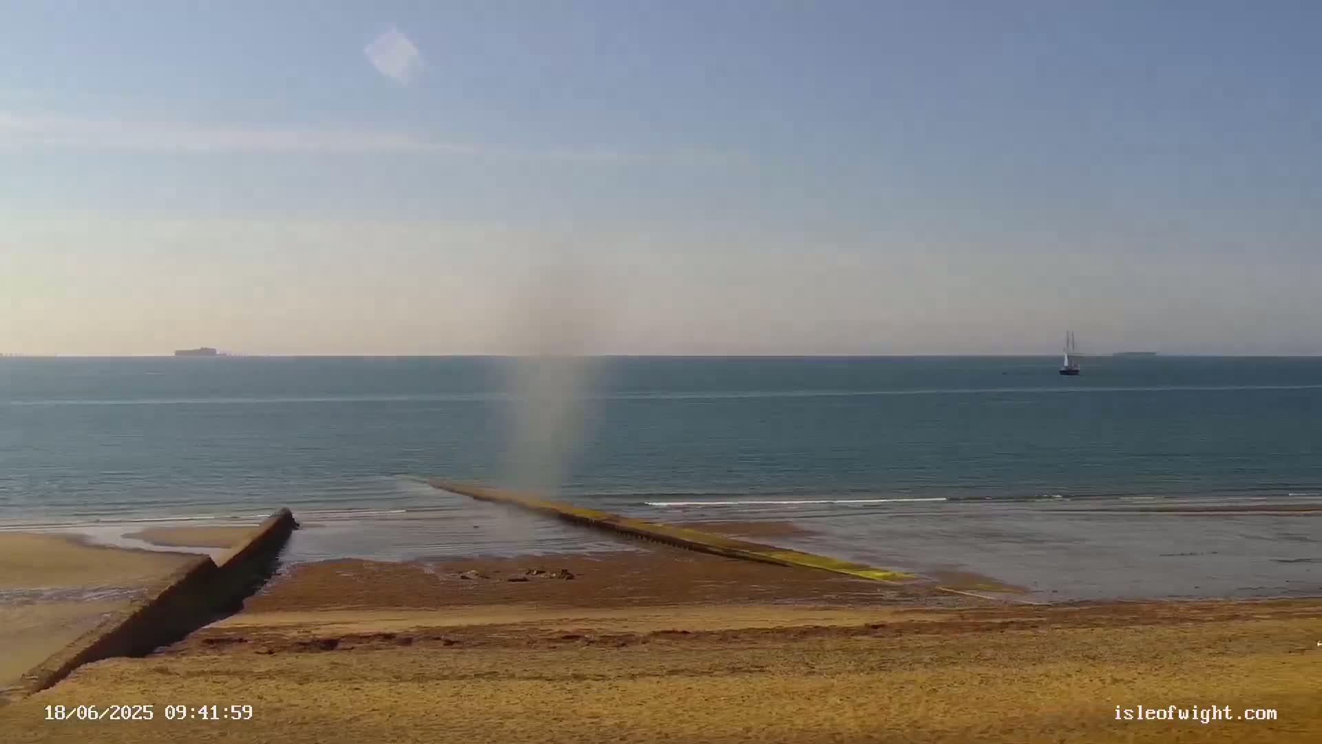 A small waterspout forms over a calm sea near a sandy beach with a long breakwater extending into the water.