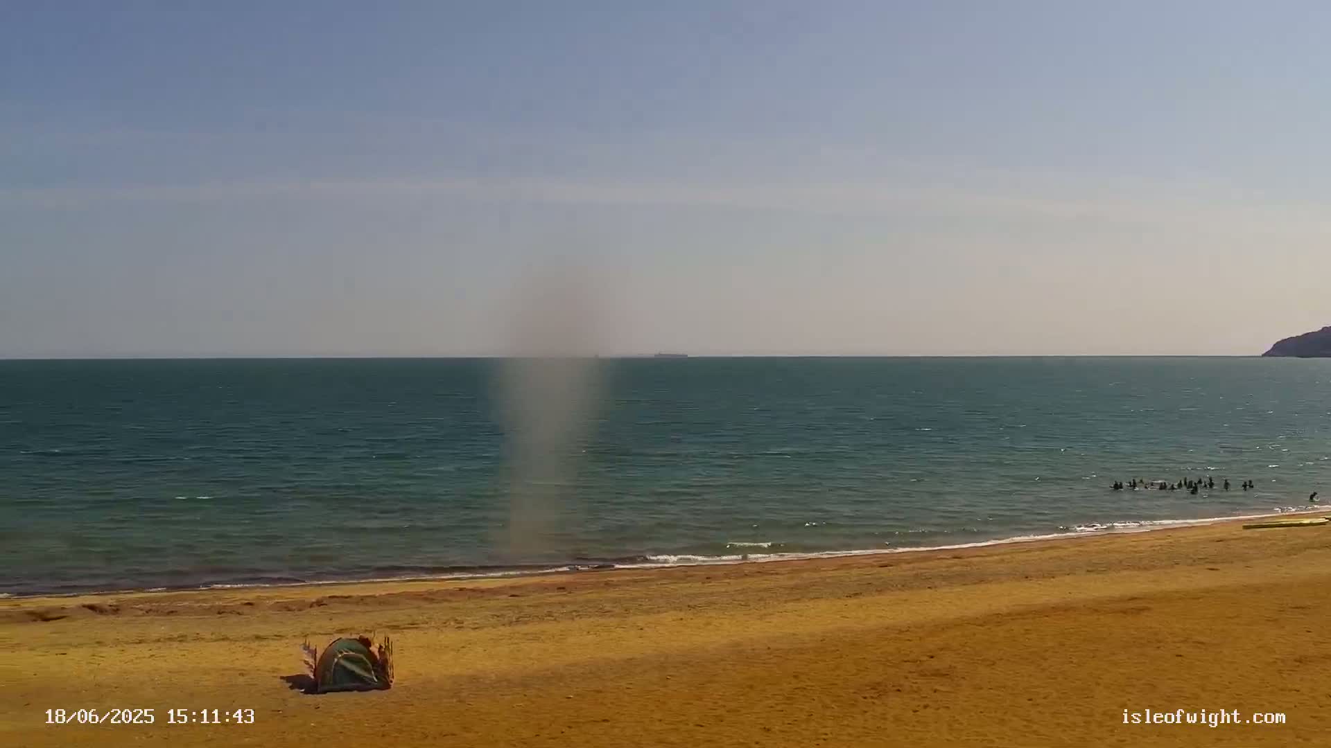 A sandy beach with a small, green tent and a group of people in the water is visible under a mostly clear sky, with a faint, vertical dust devil or waterspout over the sea in the distance.