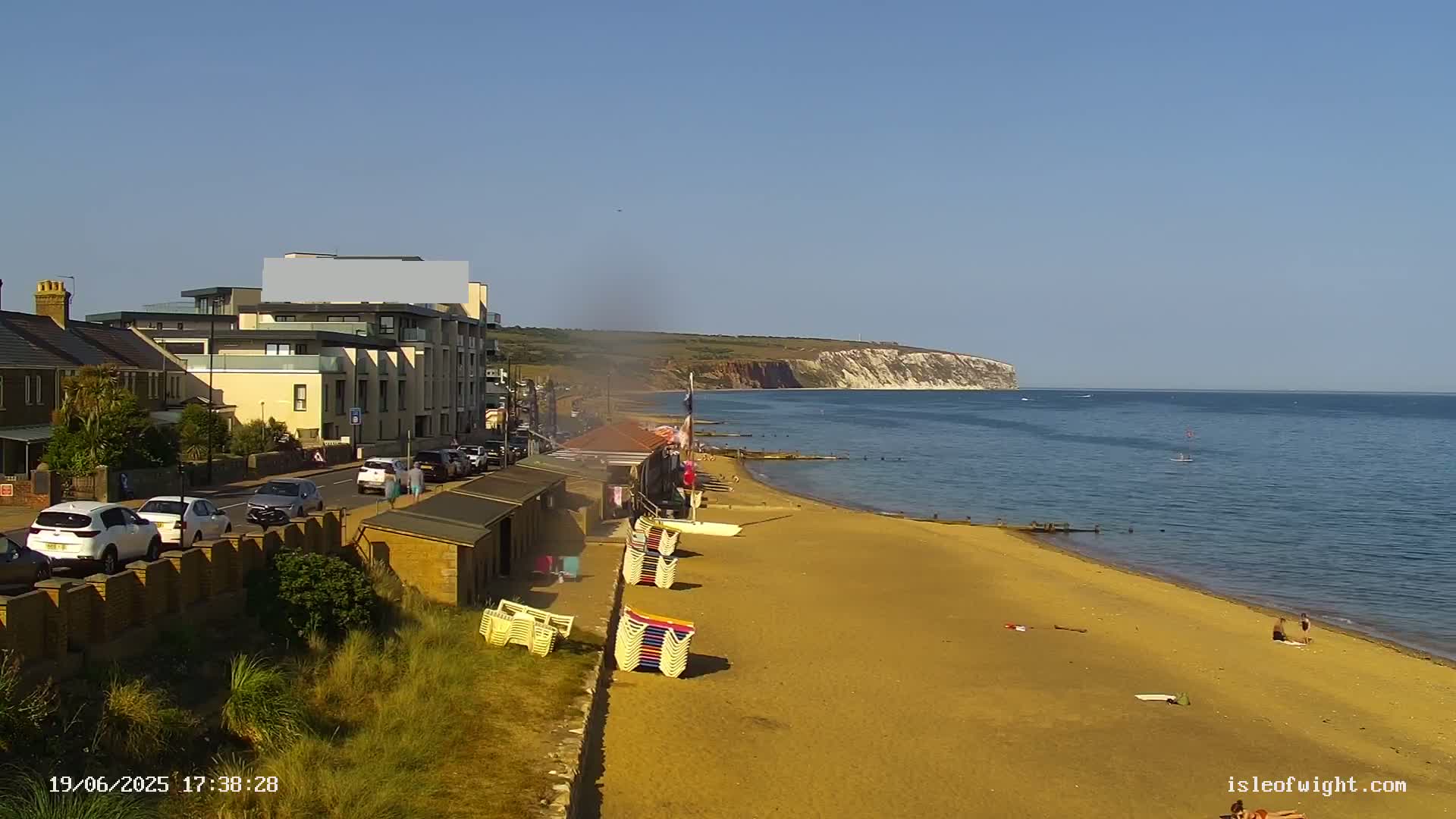 A sandy beach stretches along a calm sea, with a seaside town, buildings, and stacked beach chairs visible in the foreground, under a clear blue sky.