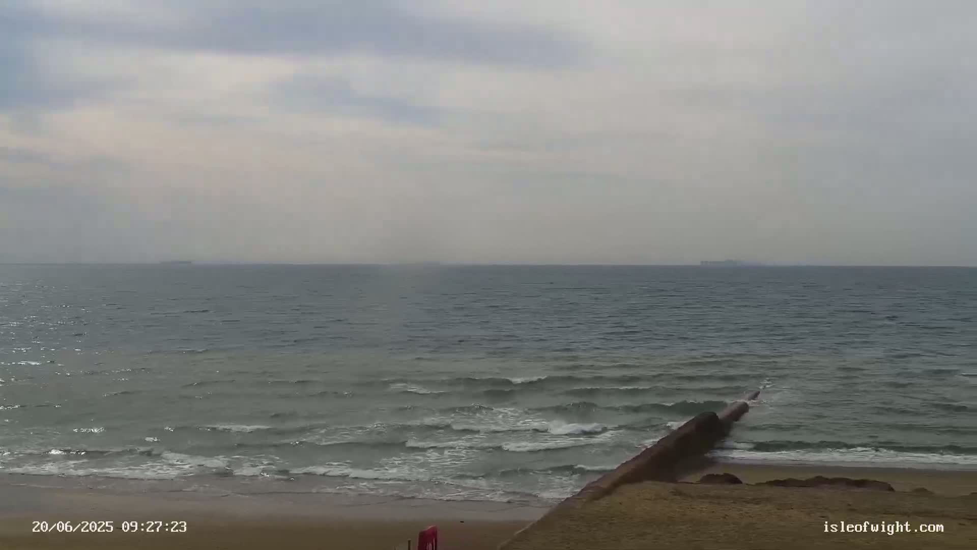 A sandy beach with gentle waves rolls onto the shore under an overcast sky, with a distant view of the sea and a few ships on the horizon.