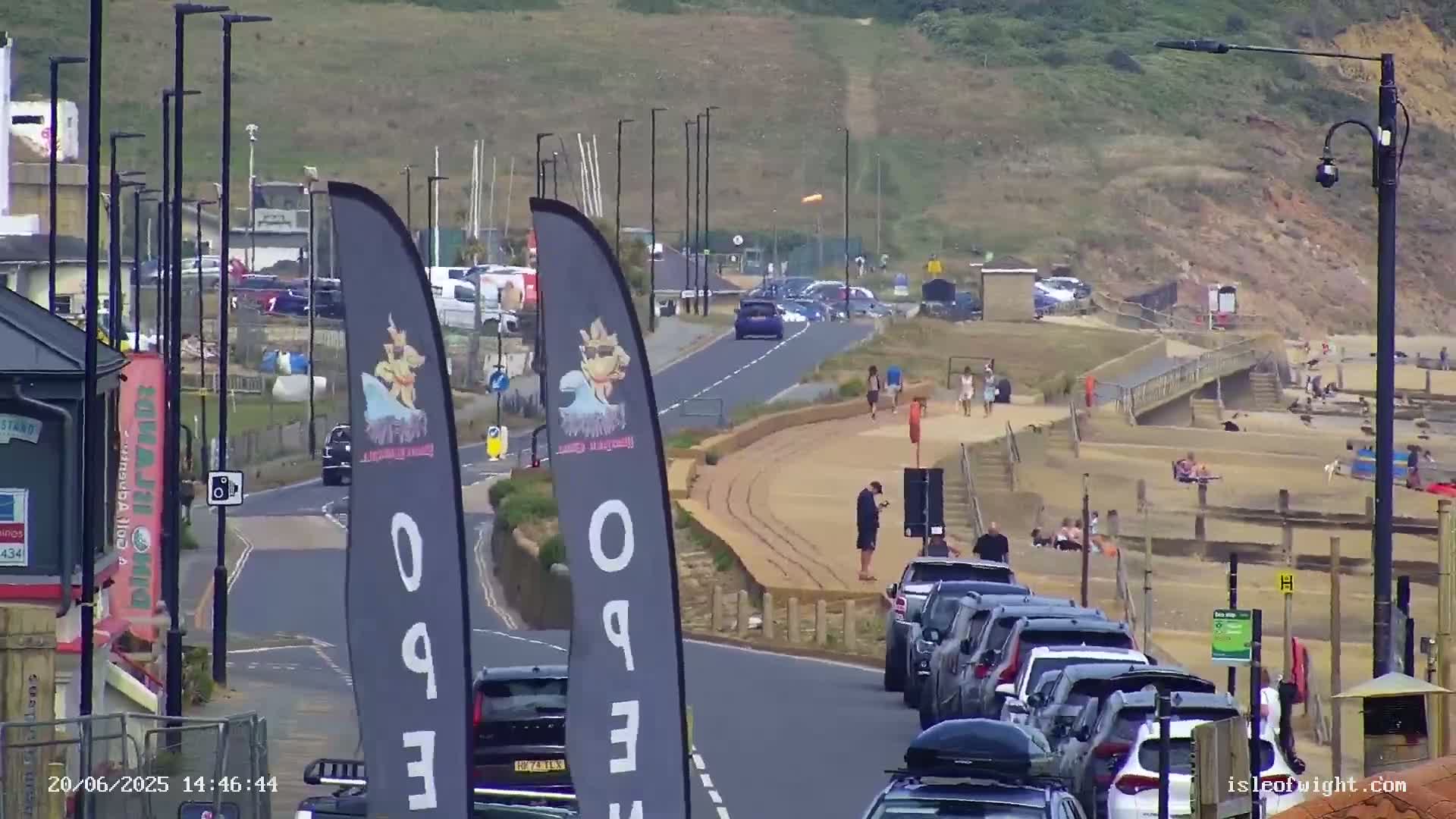 A sunny seaside road with parked cars and pedestrians on a sandy beach.