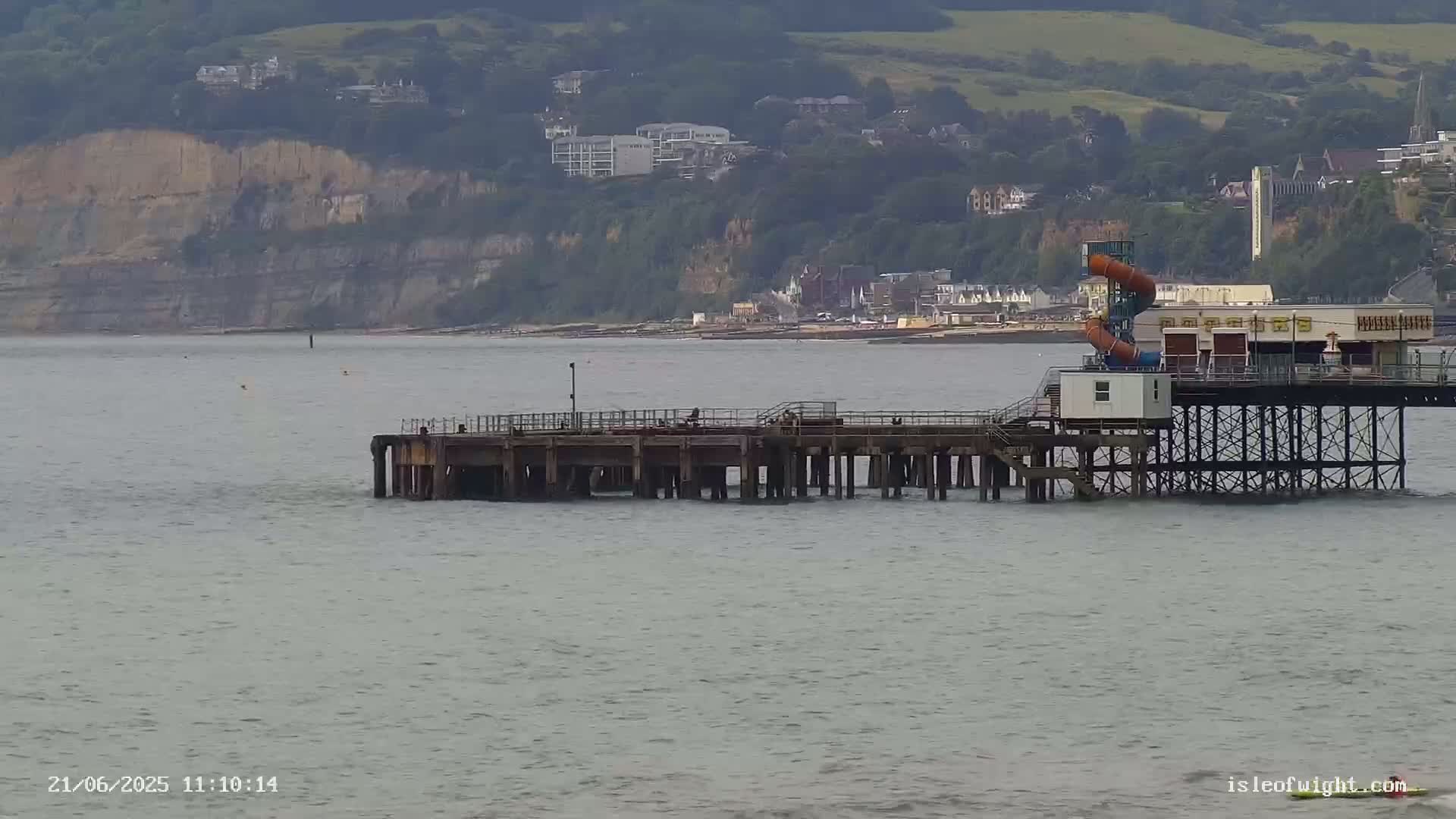 A pier extends into a calm, gray sea, with a water slide visible on the pier and cliffs and buildings in the background.