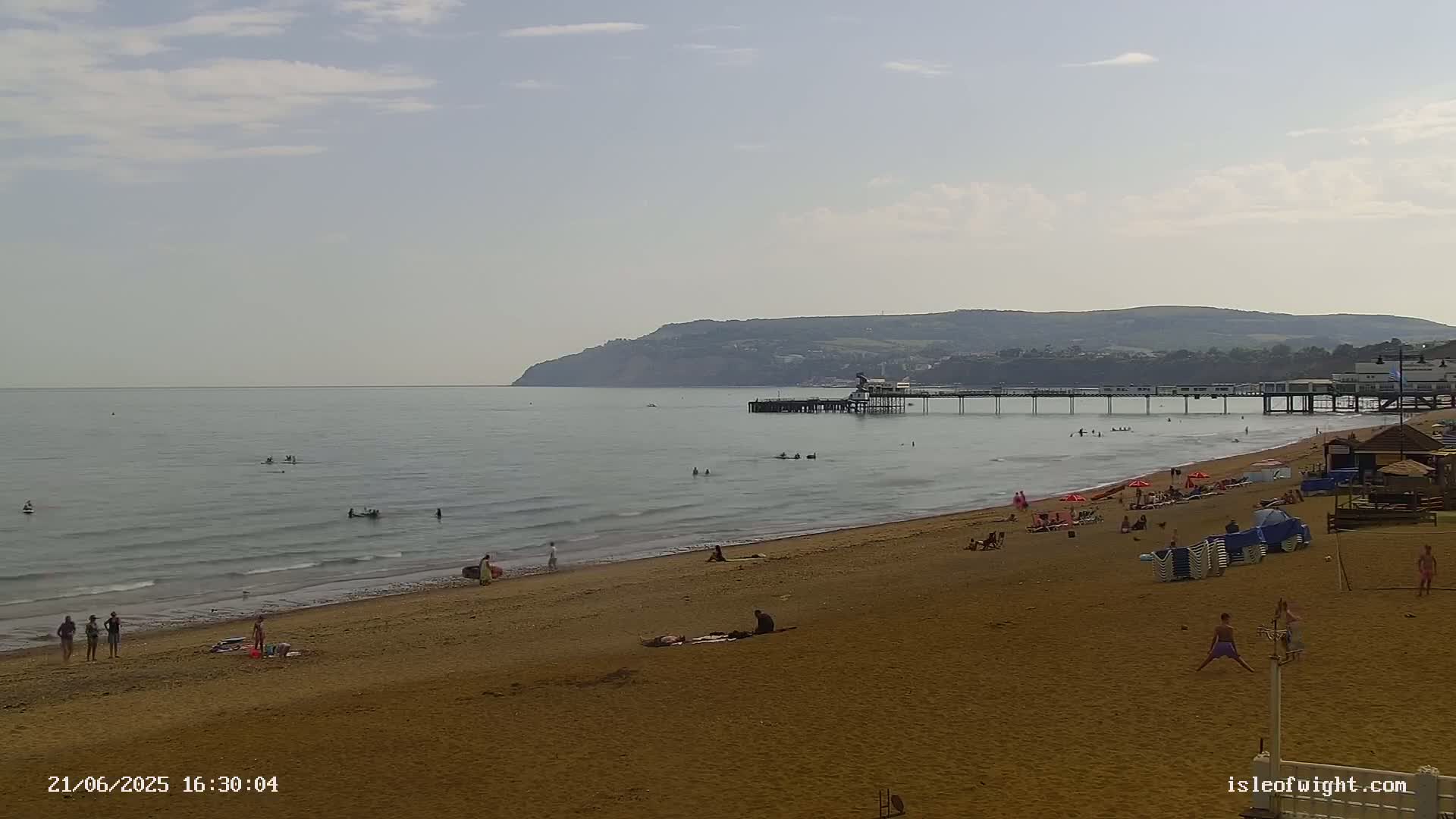 A sandy beach with people swimming, sunbathing, and playing, a pier extending into a calm sea under a mostly clear sky, and a hill in the background.