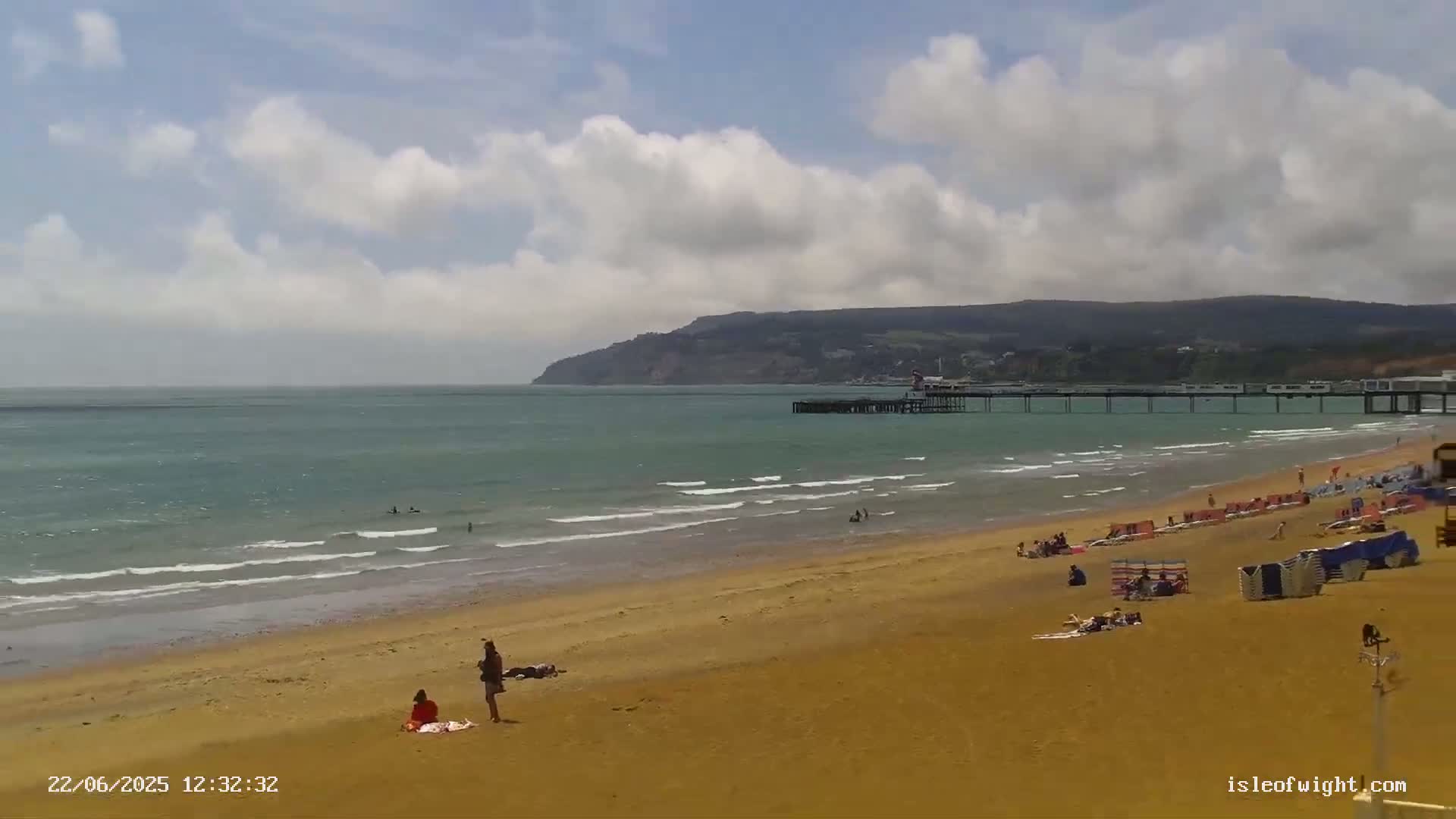 A sandy beach with people sunbathing and swimming, a long pier extending into a calm sea under a partly cloudy sky, and a large hill in the distance.