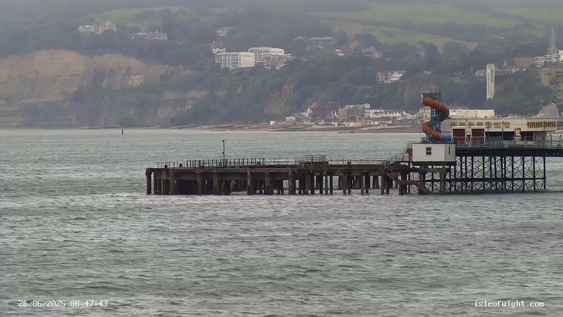 A pier with a water slide extends into a choppy gray sea, with a town and cliffs visible in the background under an overcast sky.