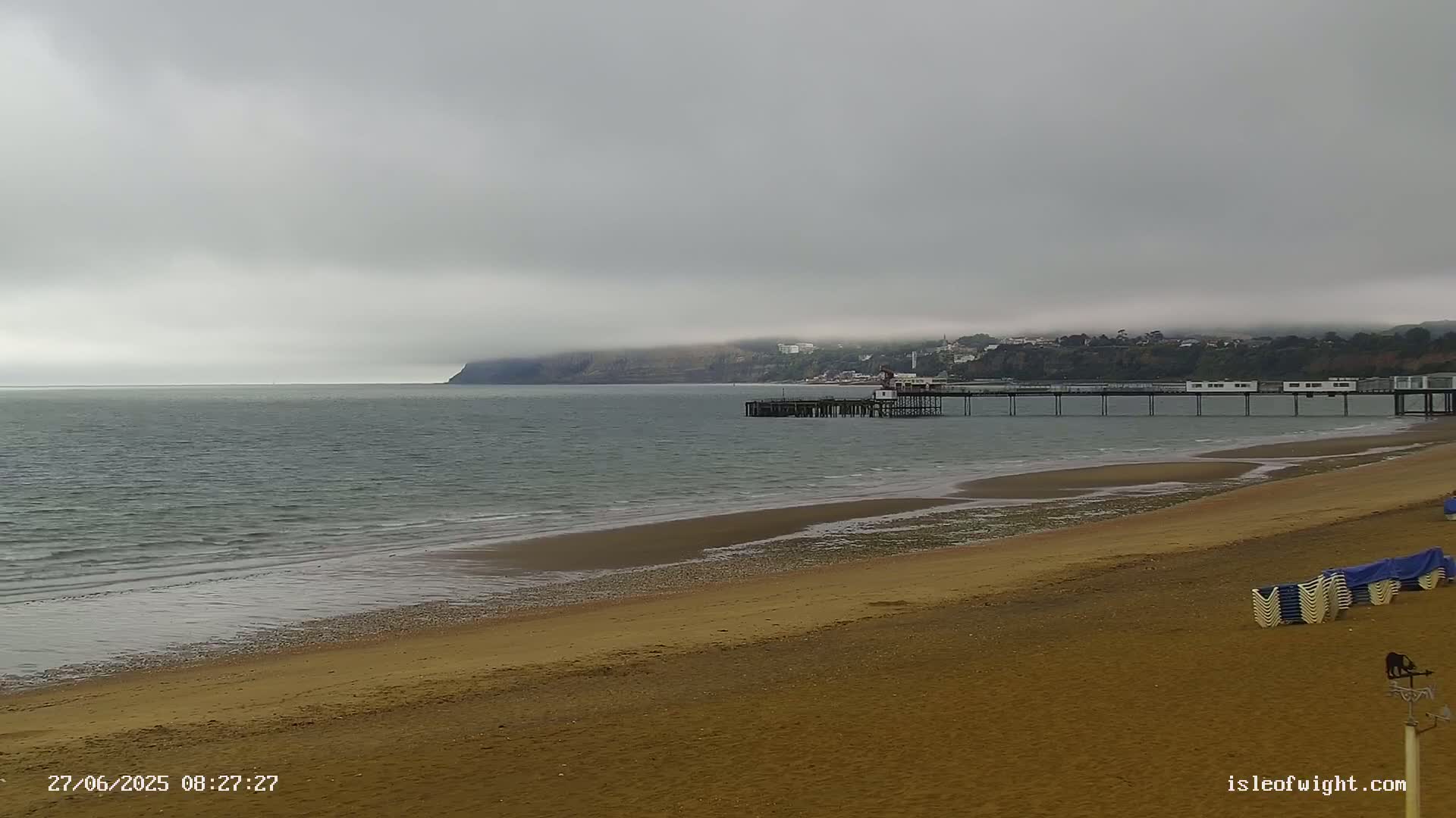 A sandy beach stretches to a calm sea under an overcast sky, with a pier extending into the water toward a distant, fog-shrouded coastline.