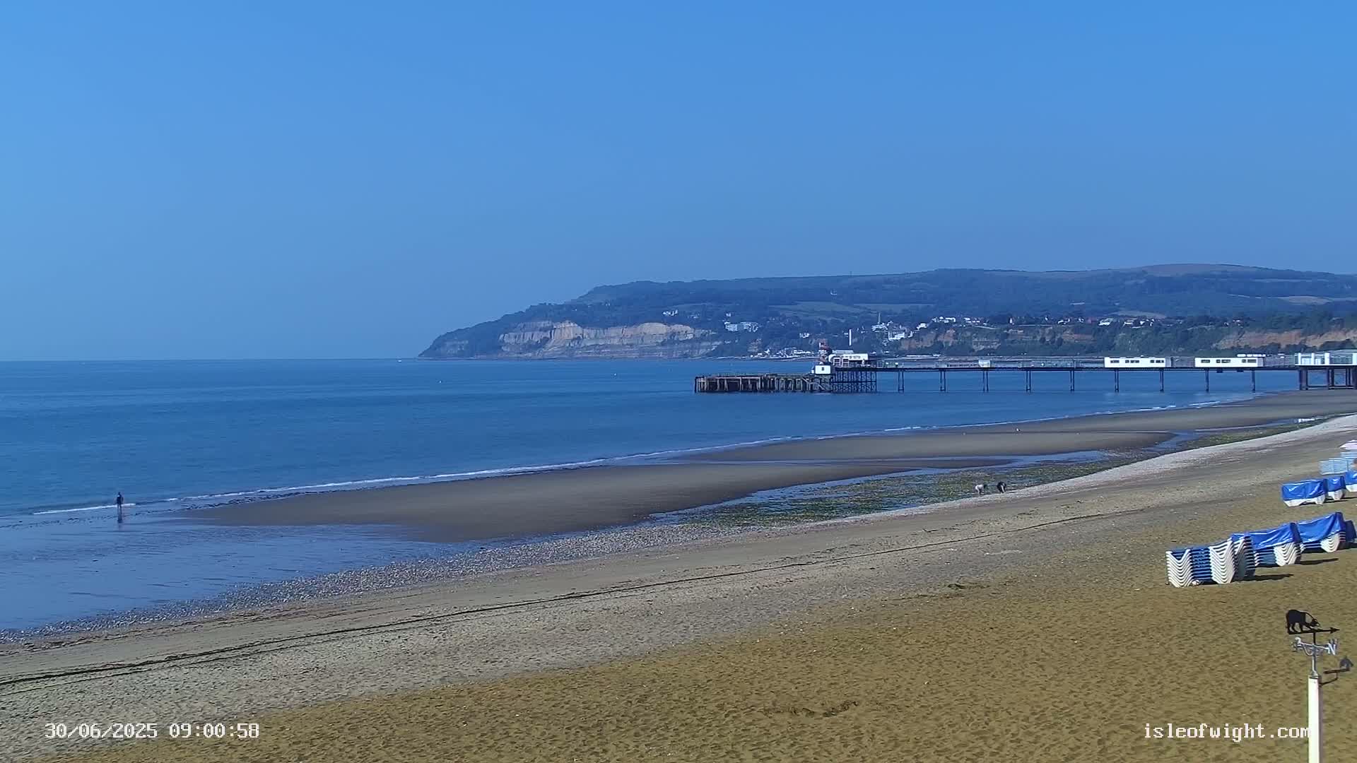 A sandy beach stretches toward a pier and calm ocean under a clear blue sky, with a distant cliff in the background.