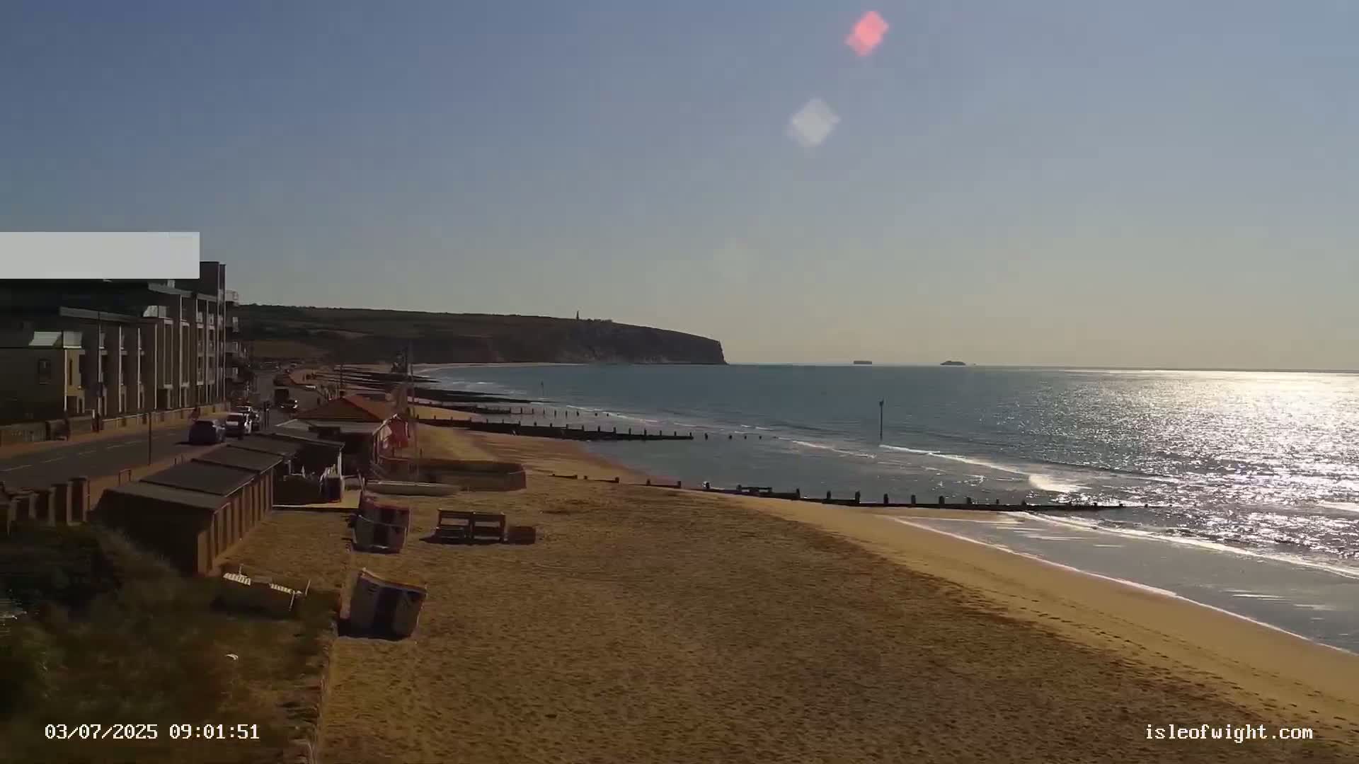 A sandy beach with small waves and calm ocean, a cliff in the distance, and a building beside a road are visible under a clear, sunny sky.