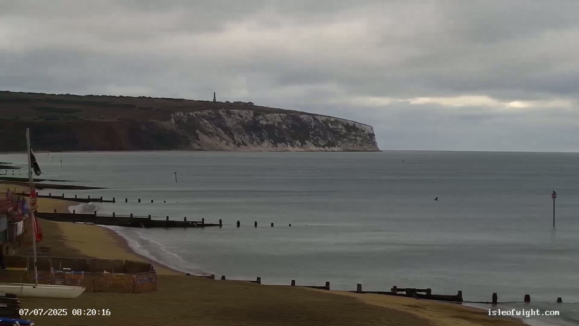 A sandy beach with calm, gray water and a distant white cliff face under an overcast sky.