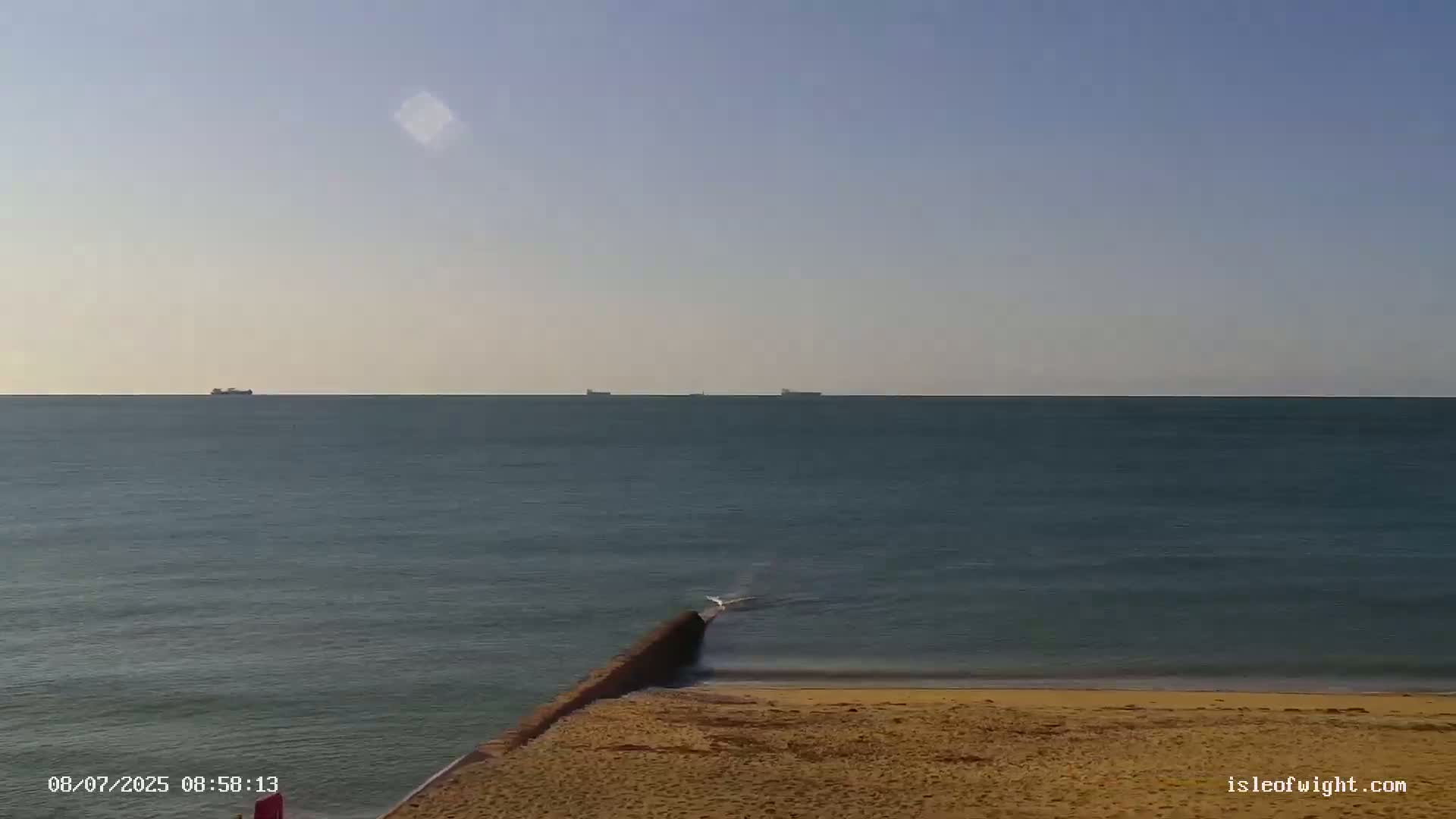 A calm sea with distant ships is viewed from a sandy beach with a low wall extending into the water under a clear sky.