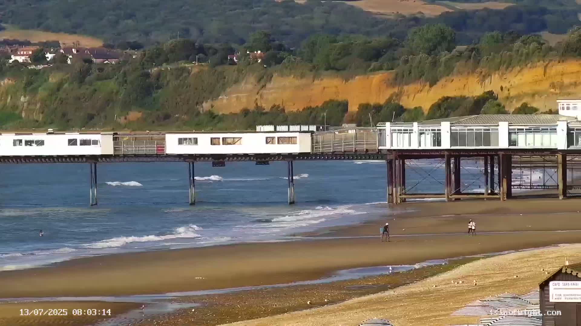 A pier with buildings extends over a calm ocean with small waves lapping a sandy beach, backed by a lush green cliff.