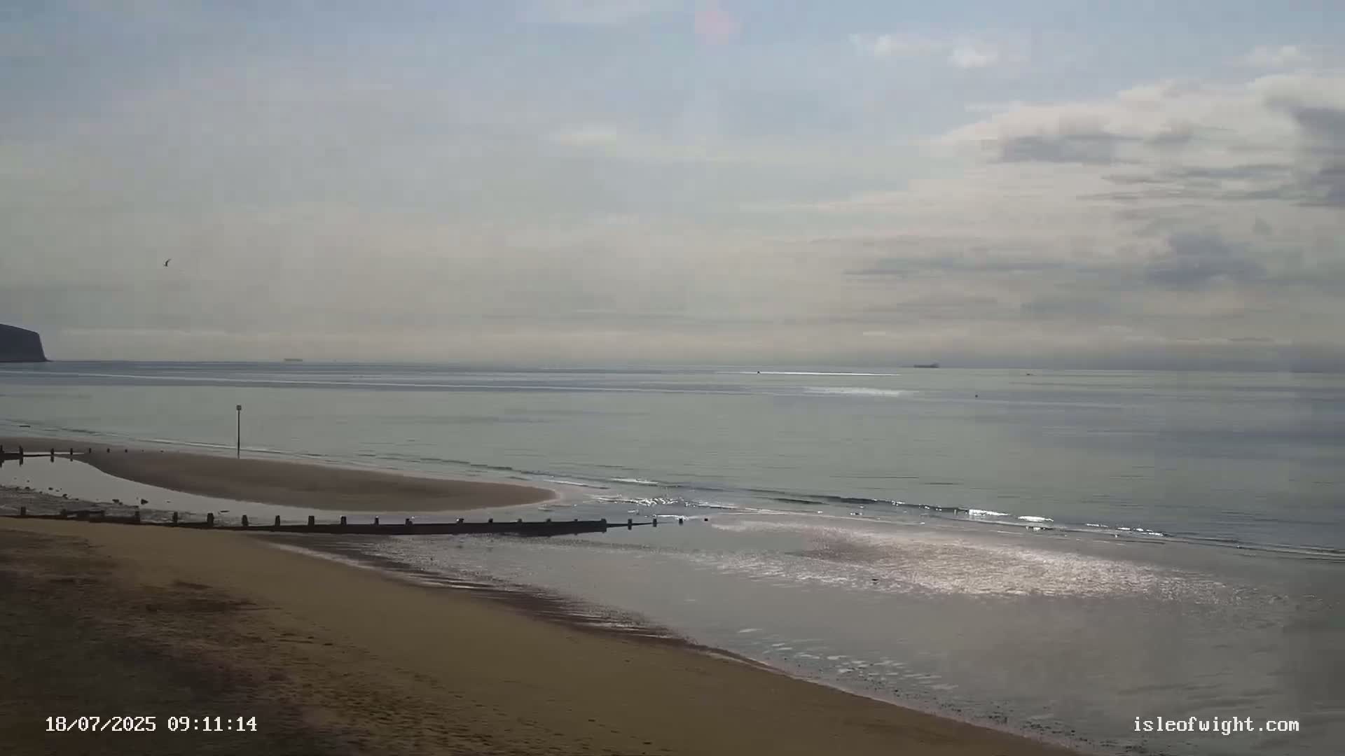 A sandy beach with calm, light-grey water under a mostly cloudy sky shows a distant cliff and a few small vessels on the horizon.