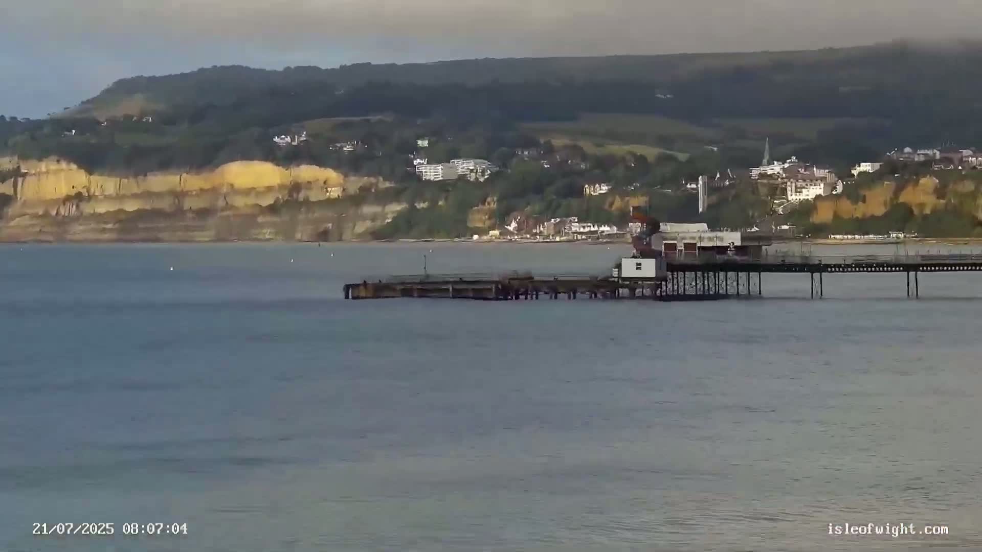 A long pier extends into a calm body of water, with a town nestled against a high, tan cliff in the background under a mostly cloudy sky.