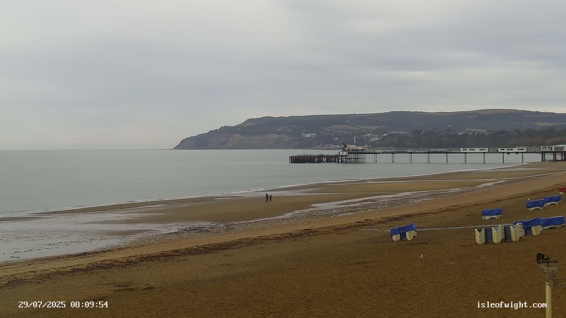 A long sandy beach stretches toward a pier extending into a calm gray sea under an overcast sky, with a distant hill visible in the background.