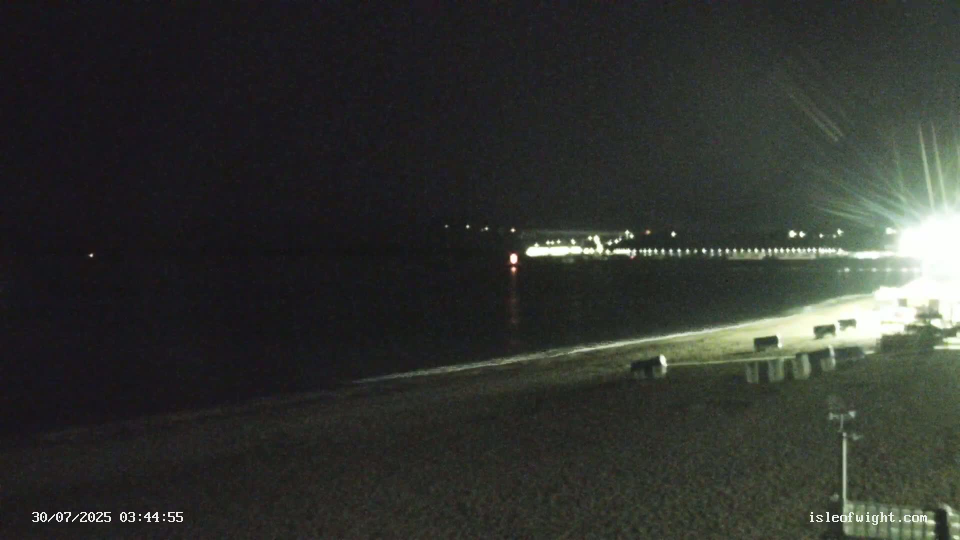 A long sandy beach stretches toward a pier extending into a calm gray sea under an overcast sky, with a distant hill visible in the background.