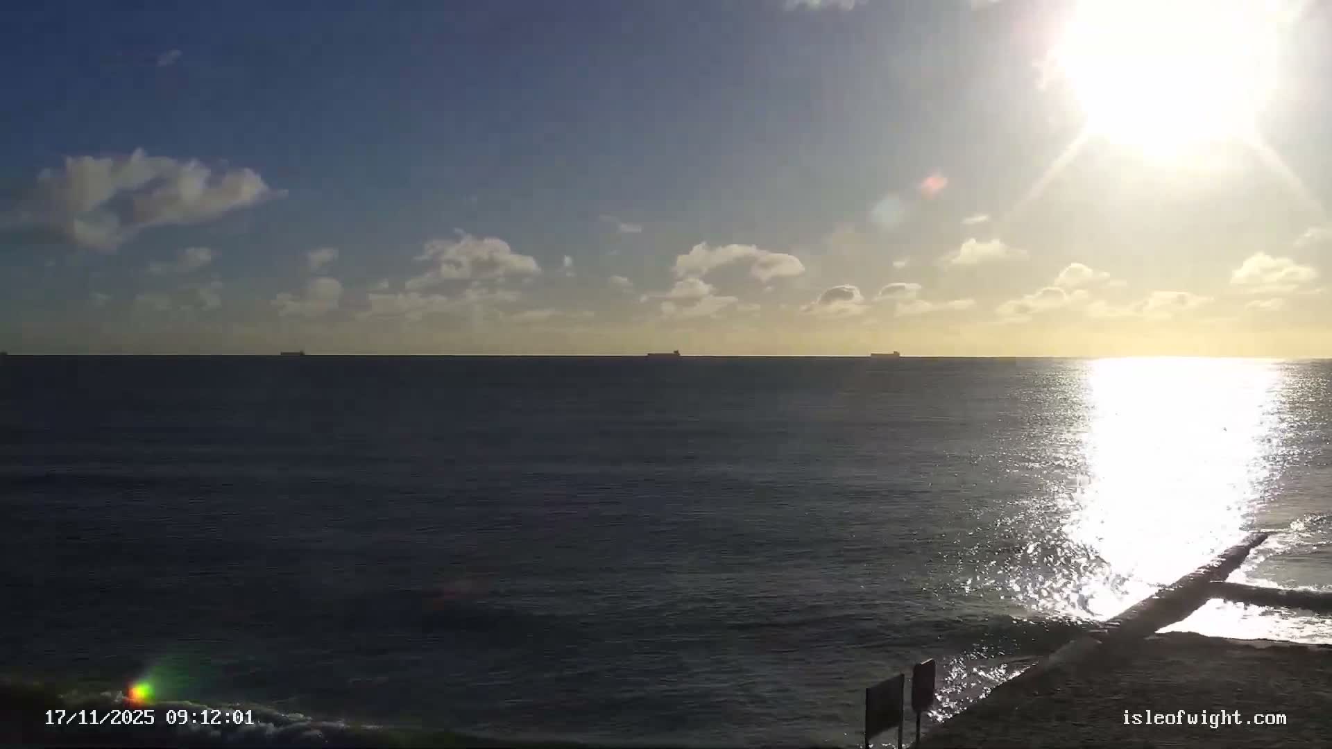 A bright, sunny day with scattered clouds illuminates a calm sea, featuring several cargo ships on the horizon and a concrete structure extending into the water on the right, all under a prominent sun glare.