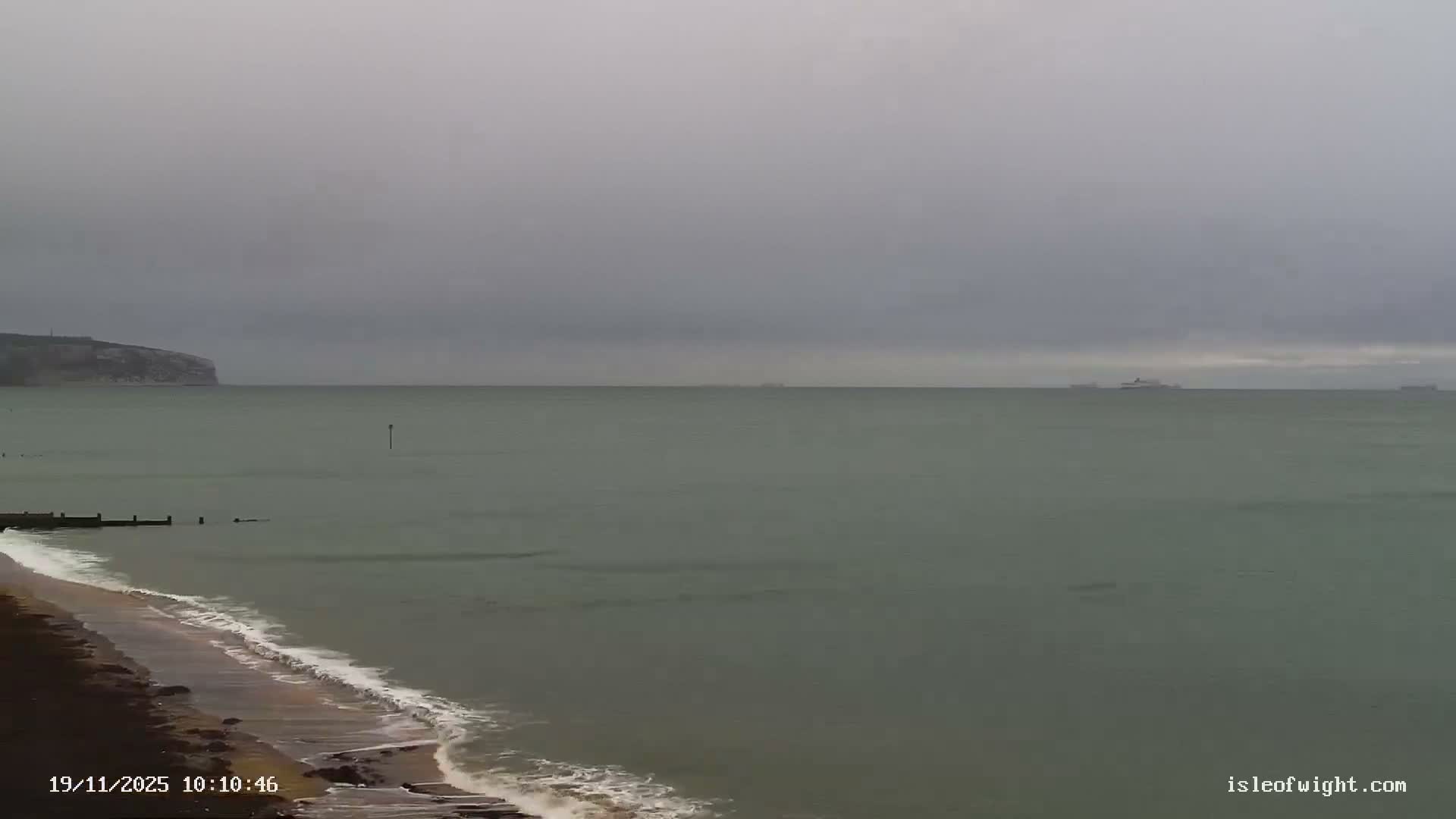 A grey, overcast sky hangs over a choppy, greenish-grey sea with waves breaking on a dark pebble beach, a distant cliff on the left, and several large ships visible on the hazy horizon.