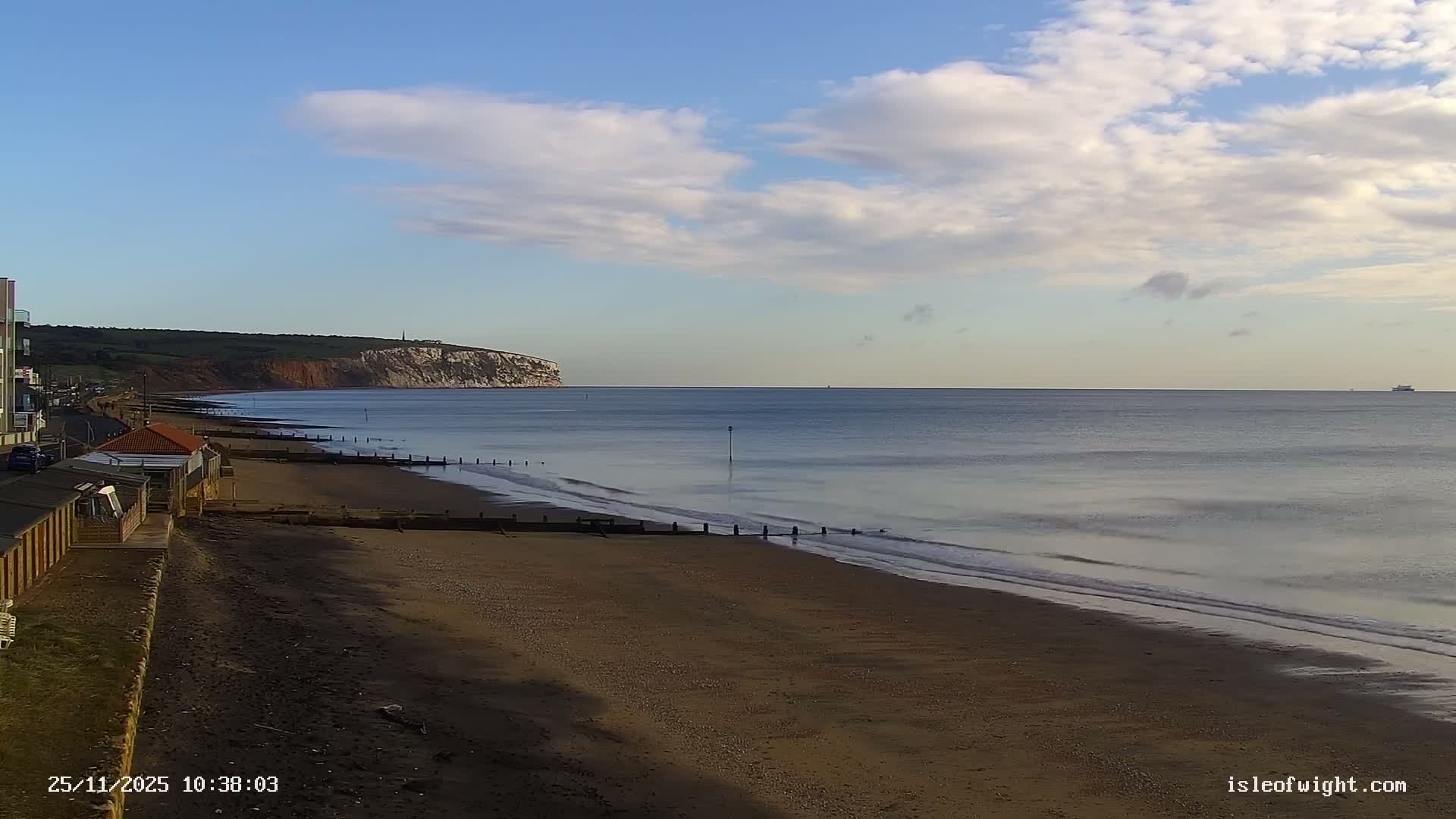 A wide sandy beach with wooden groynes meets calm blue sea under a partly cloudy sky, with a long white cliff and green hills stretching into the distance and a ship on the horizon, all bathed in bright sunlight.