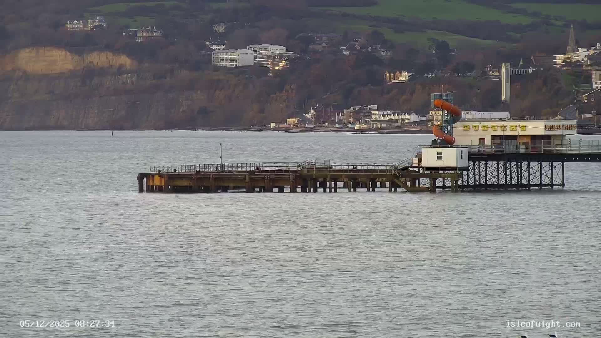 A long pier with a building featuring orange water slides extends over calm, grey water towards a coastline lined with homes and cliffs under an overcast sky.