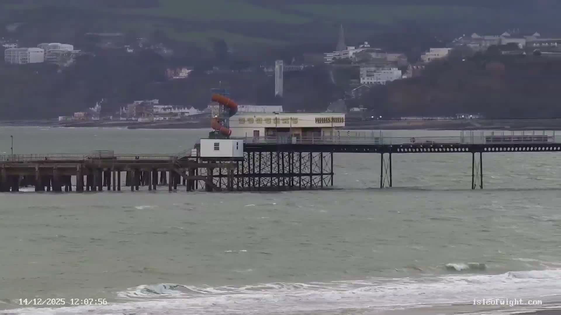 A long pier with a building featuring orange water slides extends over calm, grey water towards a coastline lined with homes and cliffs under an overcast sky.