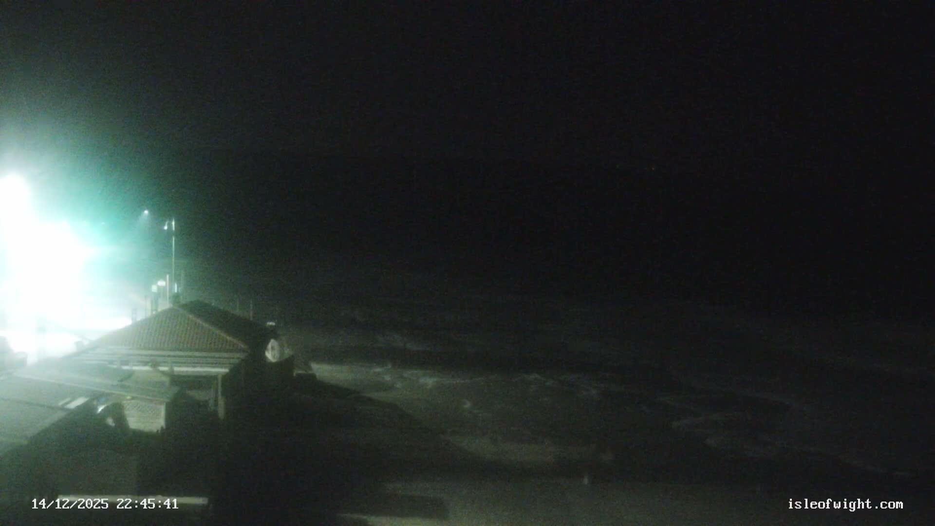 A long pier with a building featuring orange water slides extends over calm, grey water towards a coastline lined with homes and cliffs under an overcast sky.