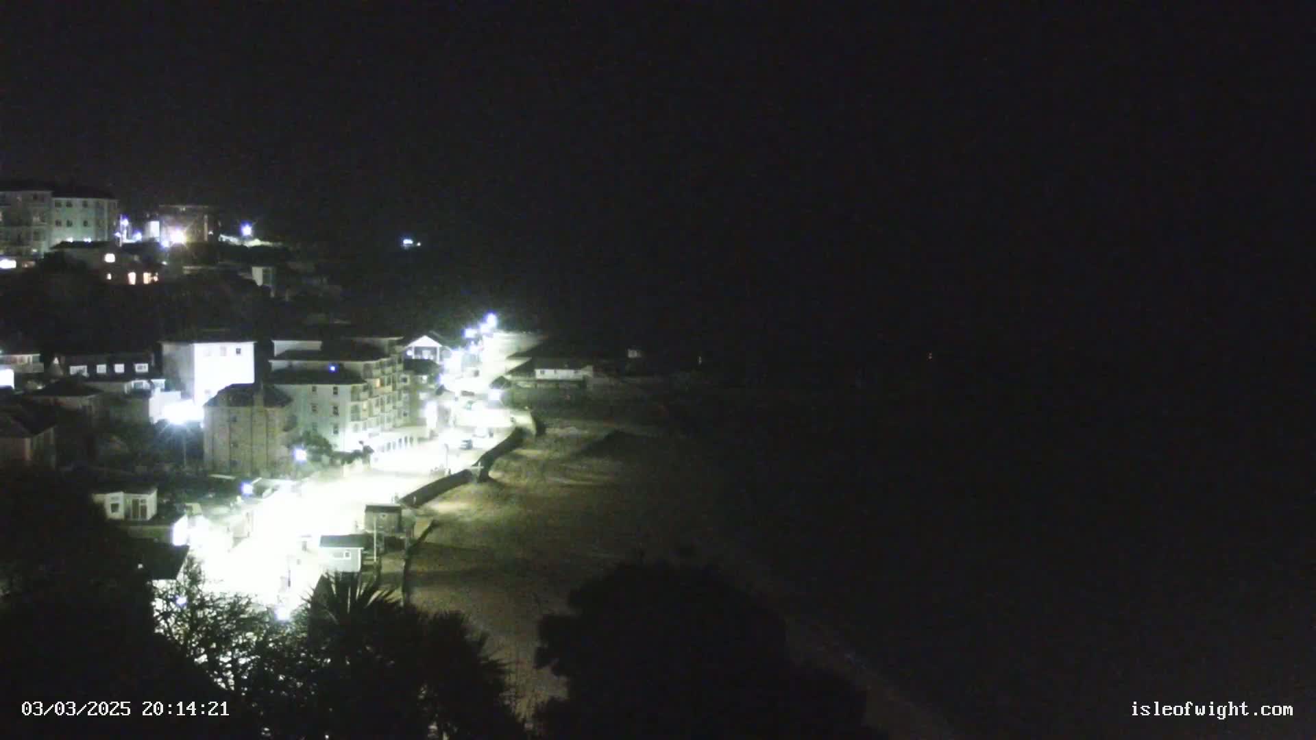 A nighttime view of a coastal town with buildings illuminated by lights, next to a dark sandy beach and a very dark ocean under a clear, dark sky.