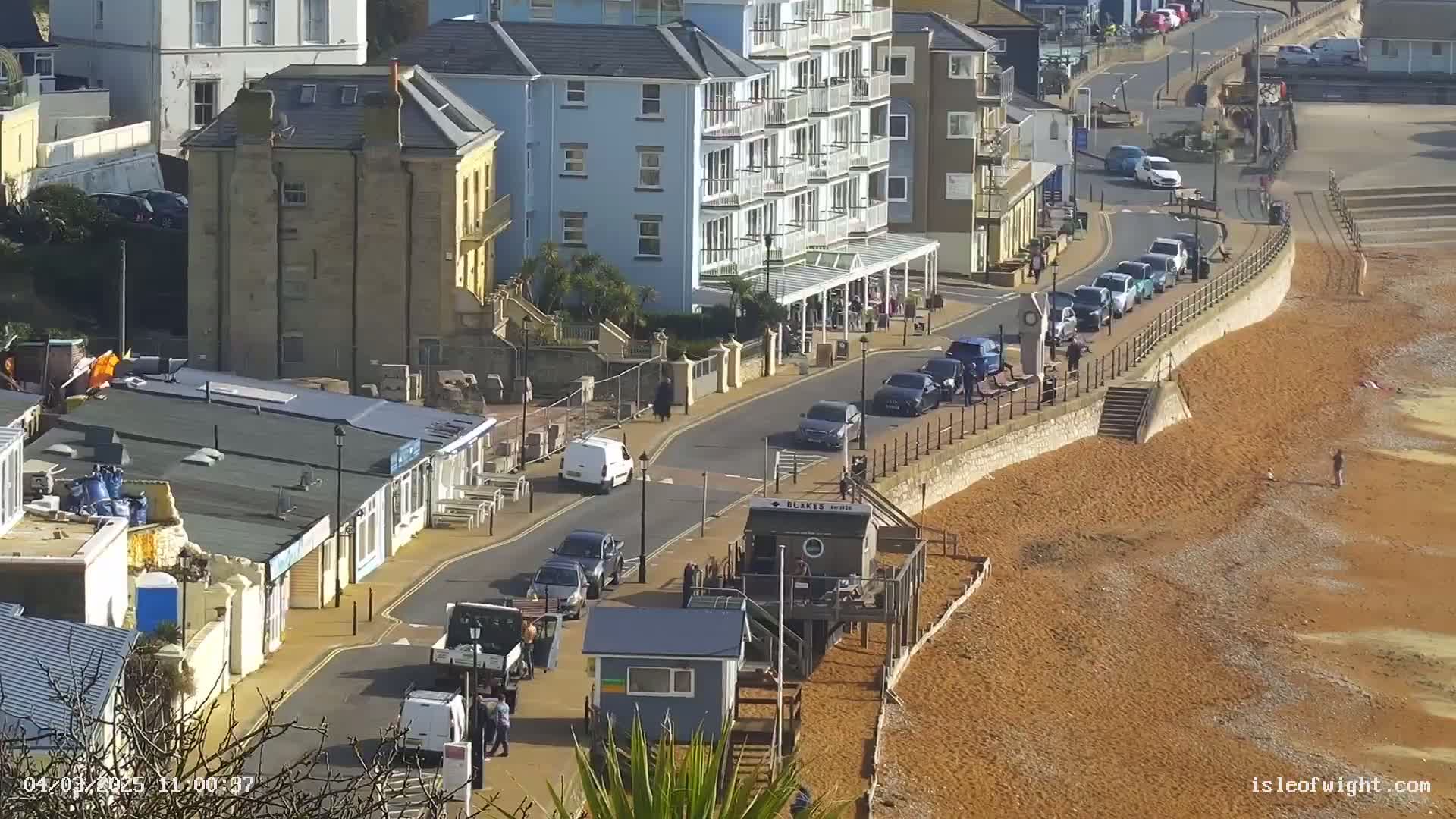A sunny seaside town street scene shows cars parked along a wall above a reddish-brown pebbled beach.