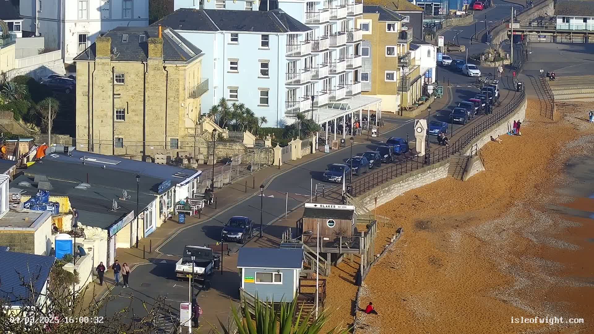 A sunny seaside town scene shows parked cars lining a road next to a sandy beach, with several buildings including a multi-story hotel and smaller shops.