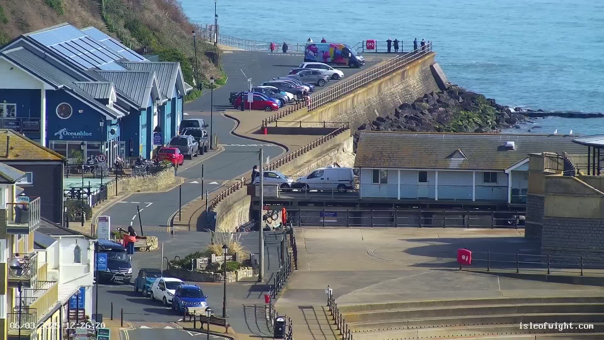 A coastal town scene on a sunny day shows buildings, cars parked along a seaside road, and a rocky shoreline with calm ocean waters.
